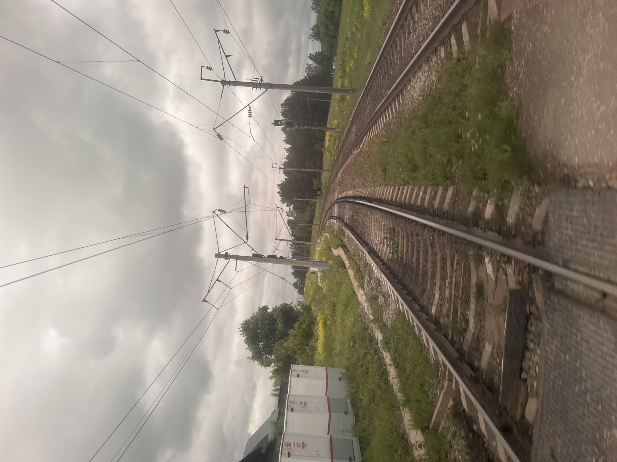 The image, originally rotated 90 degrees clockwise, depicts a railway scene under an overcast sky in what appears to be a rural or semi-rural setting in Пычас, Russia.
Two sets of parallel railway tracks curve gently to the right, receding into the distance. The tracks are laid on gravel ballast with visible wooden sleepers. Overhead electrical lines, part of a catenary system, extend across the sky, supported by multiple tall utility poles equipped with insulators and crossbeams. A green railway signal light is visible on one of these poles in the mid-ground.
Grassy embankments, interspersed with some yellow wildflowers, flank the tracks. Beyond the tracks and grass, a dense line of trees forms a forested area in the background. On the right side, parallel to the tracks, there is a concrete or asphalt platform or walkway with a textured surface. In the foreground on the left, several large, white rectangular units, possibly electrical cabinets or modular buildings, are stacked two high, featuring subtle red stripe details.
No specific activity or event is captured; the scene is static, portraying the railway environment. The sky is heavily overcast with dense grey clouds, indicating a dull, possibly damp or pre-stormy, daytime. The overall atmosphere is muted. No legible text is visible on any objects in the image.