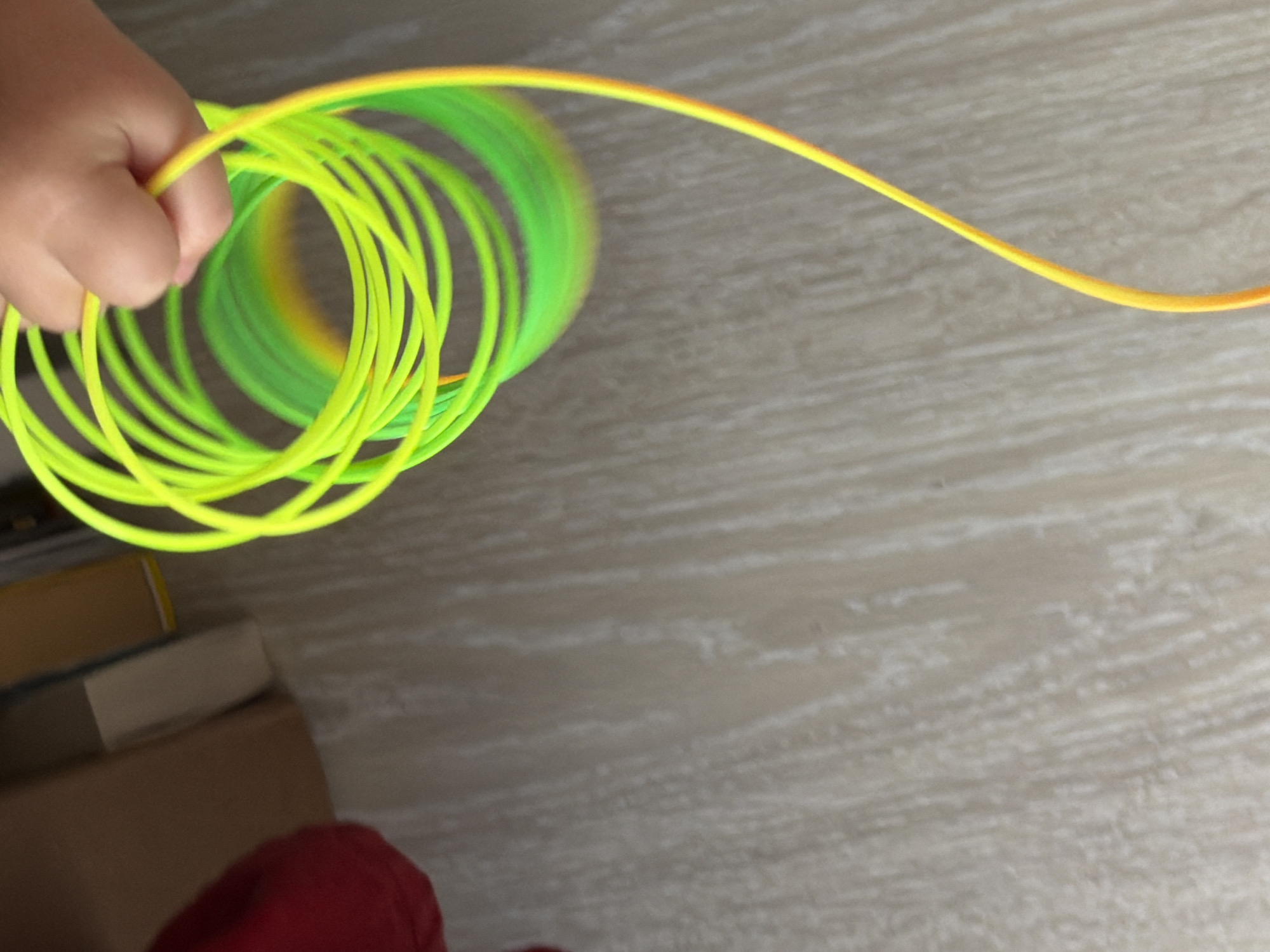 The image presents a close-up, high-angle view of a child's hand holding a vibrant, coiled object indoors in Chaikovsky, Russia. The hand, visible in the upper left of the frame, is light-skinned and grips several loops of a flexible, tube-like material. This object is predominantly fluorescent yellow-green, with sections varying in hue, appearing more green in the inner coils and transitioning to a brighter yellow and subtle orange along its length. It forms multiple concentric circles, suggesting it is either a spring-like toy, a hula hoop, or a length of rope being manipulated.
The background consists mainly of a light-colored, textured surface, resembling a wooden floor or laminate, with discernible grain patterns. In the bottom left corner, partially visible, are stacked items that appear to be books or cardboard boxes, one with a brown hue and another with a darker spine. A small patch of red fabric, likely part of clothing, is also visible at the very bottom. The activity involves the hand actively holding or playing with the coiled object. There is no visible text in the image, and no specific time of day or emotional state can be inferred. The lighting suggests an indoor setting with ambient illumination.