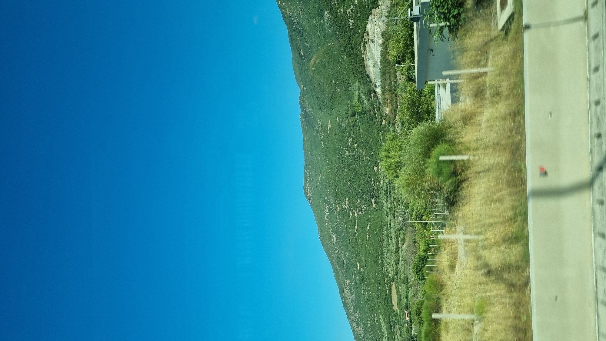 The image captures a natural landscape under a clear, vibrant blue sky. In the background, a lush green mountain slopes upwards, covered in dense vegetation. The lower portion of the image shows a dry, grassy field with scattered bushes and a fence line. To the right, a small, modern-looking building with a grey facade is partially visible, situated behind the fence. A concrete barrier runs along the bottom of the frame, suggesting a roadside perspective. The scene is set in the Municipal Unit of Ampelakia, Greece.