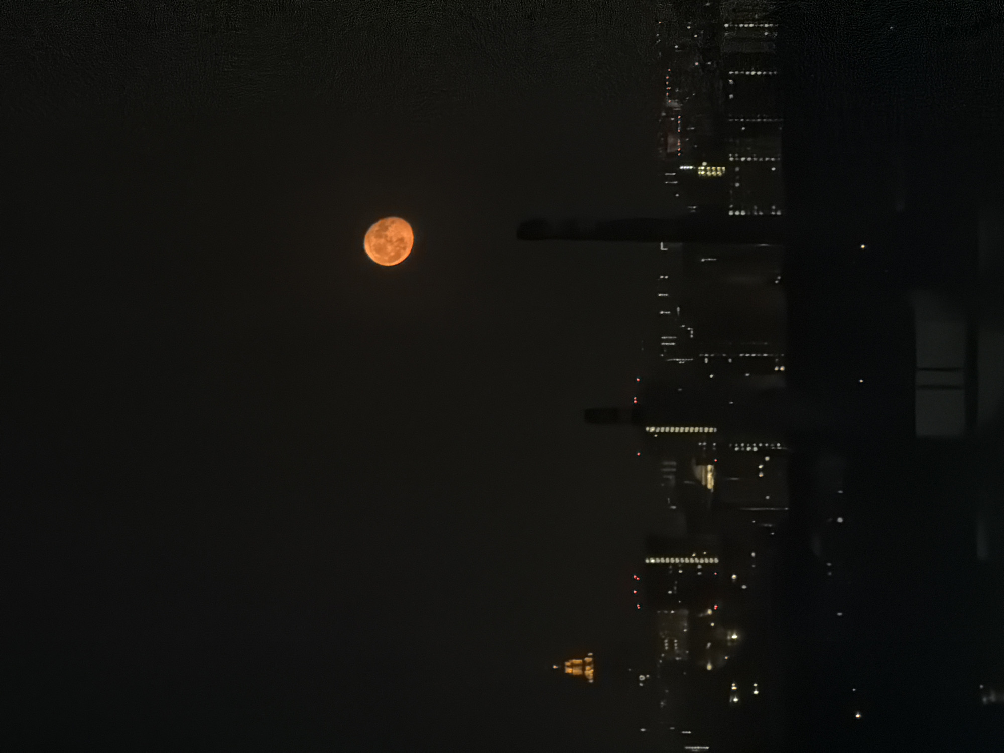 The image depicts a full moon in a dark sky, observed from a high vantage point, likely a tall building. The scene includes the night cityscape, reflected in the foreground, showing the blurred outlines of buildings and scattered lights. The reflection suggests the photo was taken through a window. The location is stated to be Khimki, Russia, but there are no elements in the image definitively confirming this location, the night cityscape is more generic. There are no people depicted.