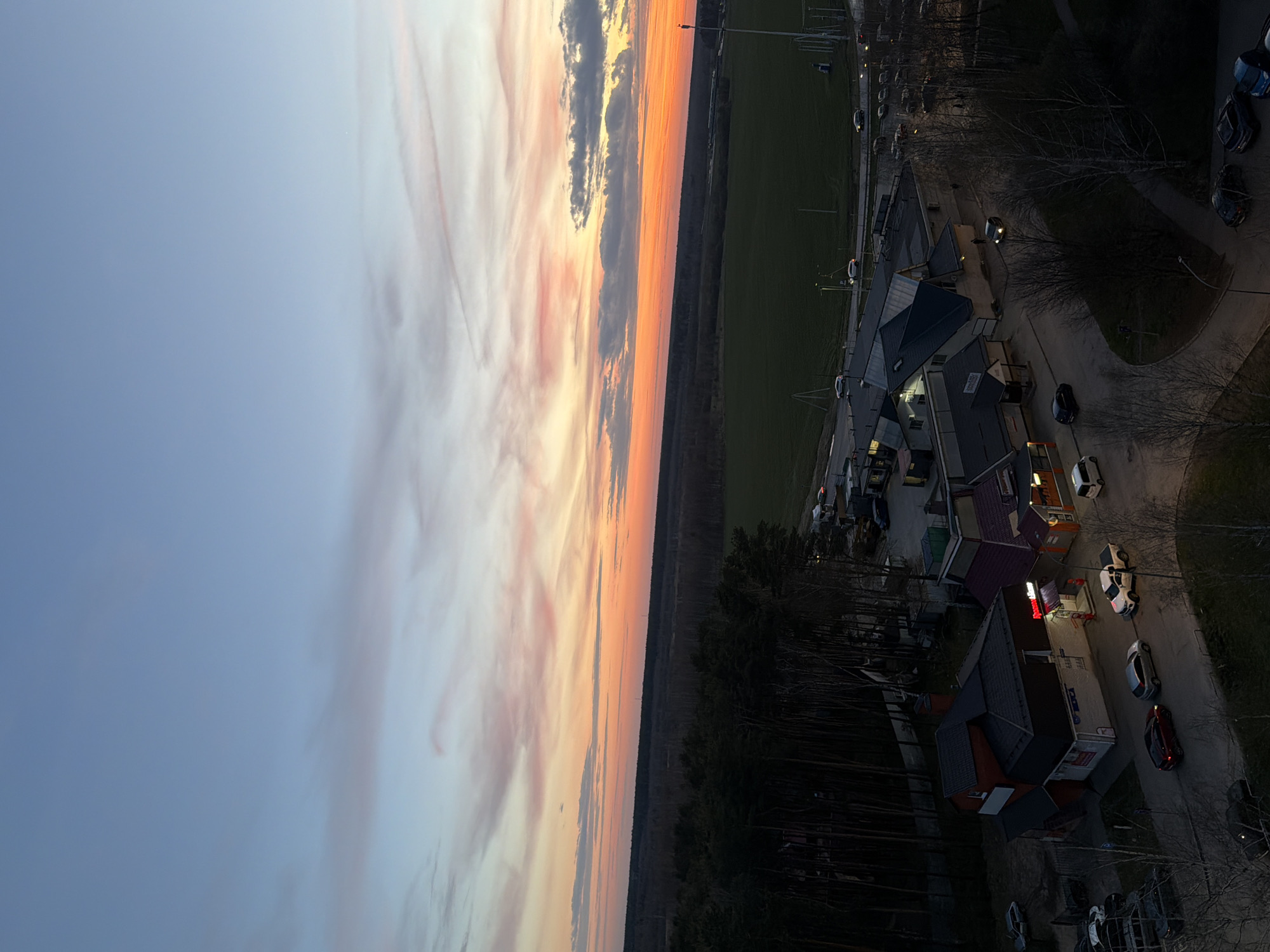 The image depicts an aerial view of a landscape during a vibrant sunset or sunrise, likely taken from a high vantage point in or near Chernogolovka, Russia. The horizon is dominated by a colorful sky with gradients of orange, pink, and grey clouds transitioning into a light blue. Below the horizon line, a dark expanse of forest stretches across the middle ground. To the right, there are signs of urban development including buildings with lit windows, cars parked along a winding road, and a green field in the distance. The overall scene captures the transition between natural and built environments.