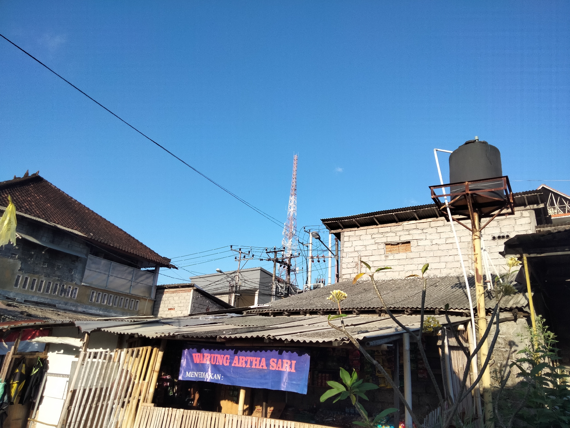 A telecommunications tower is visible in the background, situated above a dense urban landscape of buildings with corrugated metal roofs. In the foreground, a rustic structure, possibly a market stall or a small shop, displays a blue banner with the text "WARUNG ARTHA SARI MENYEDIAKAN:". A large black water tank on a metal platform is mounted on a pole above some of the buildings. The scene is illuminated by natural daylight, casting shadows from the structures and vegetation. The sky is clear and blue. The general appearance suggests a scene from a residential or commercial area in Denpasar, Indonesia, characterized by a mix of older and newer construction, and common urban infrastructure.