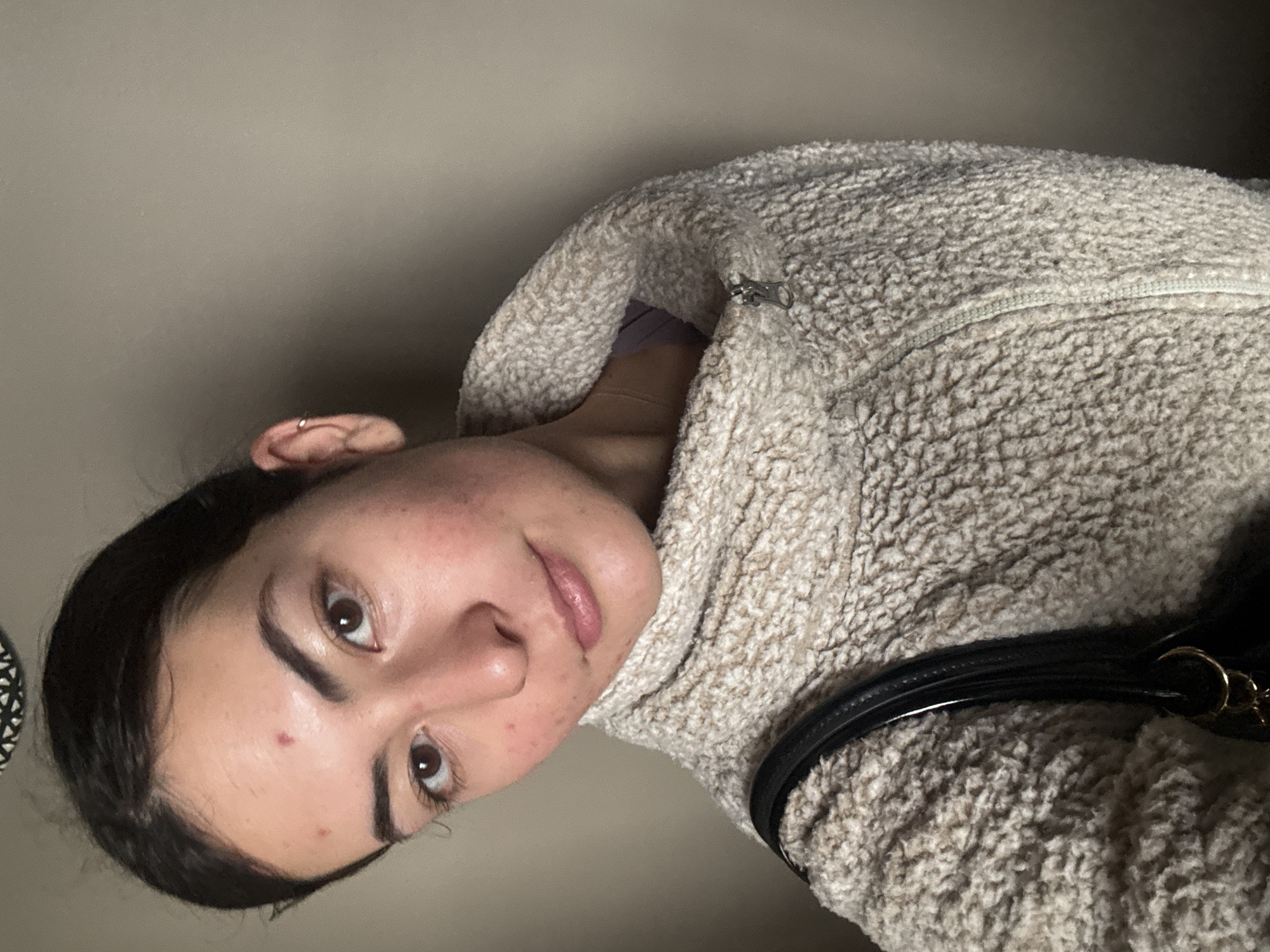 A woman with dark hair pulled back is wearing a light beige, textured fleece jacket. A black purse strap is visible over her right shoulder. She has fair skin with a few blemishes and a mole on her forehead. She is wearing a small hoop earring in her left ear. The background is a plain, light gray wall. The location is not discernible from the image.