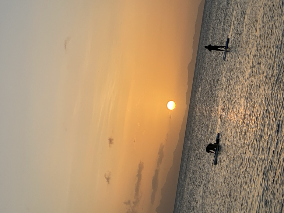 The image displays a coastal sunset scene in Juan Griego, Venezuela. The bright orange sun is prominently positioned above the horizon, casting a warm glow across the sky, which transitions from deep orange and yellow near the sun to lighter hues higher up, with minimal scattered clouds. The calm water surface, exhibiting ripples, reflects the sunset. Two individuals are present on the water, both on paddleboards. The subject on the left is kneeling on a paddleboard, while the subject on the right is standing upright on a paddleboard, holding a paddle. Both individuals are silhouetted against the bright background. A range of distant mountains forms a dark, undulating silhouette along the horizon line.
