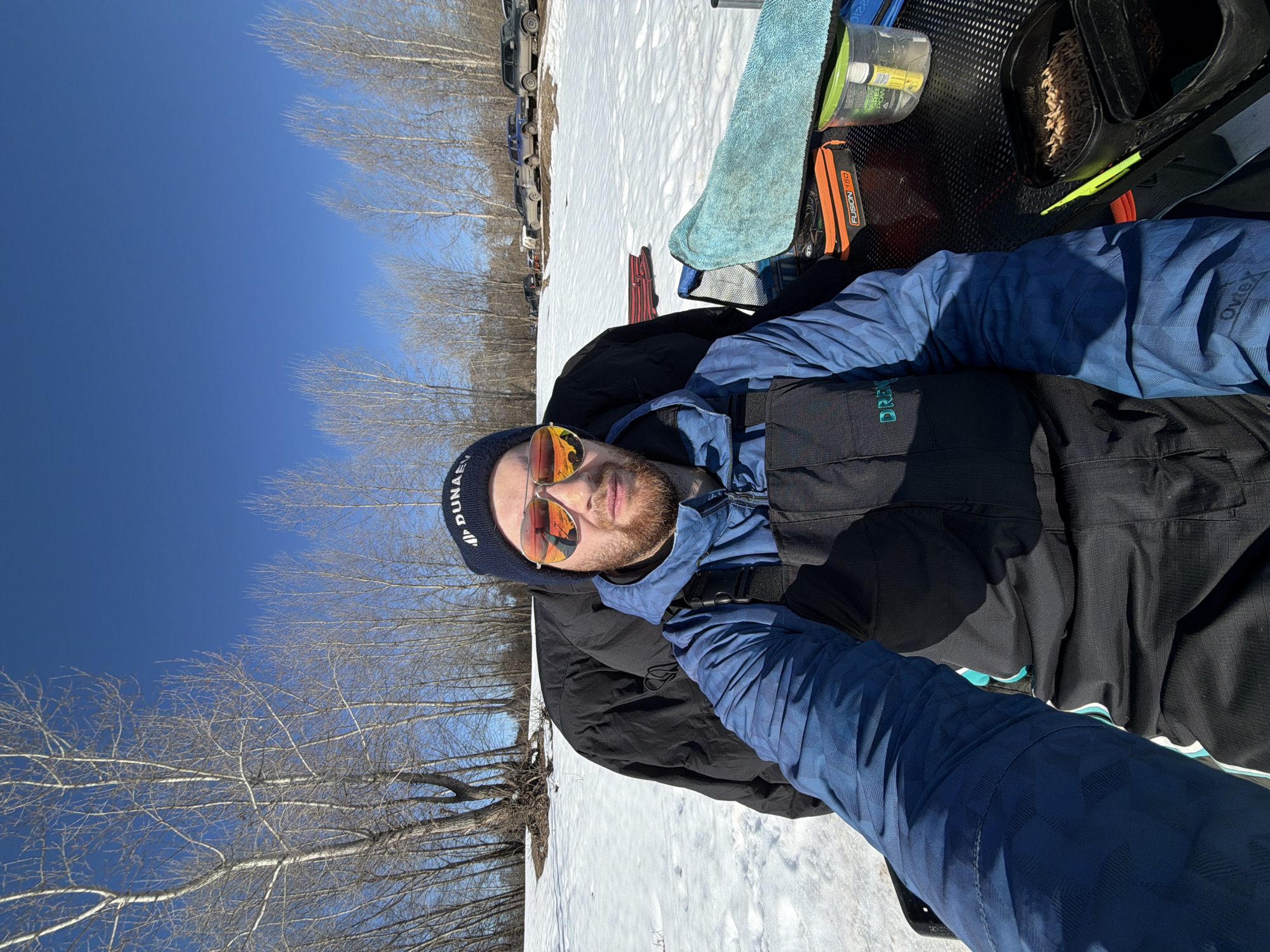 A man lies on a snow-covered surface outdoors. He is wearing a blue beanie with white text, sunglasses with orange lenses, a black vest, and a blue patterned jacket. He has a beard and fair skin.
To the left, bare trees stretch upwards against a clear blue sky. In the distance, parked vehicles are visible. The man is positioned near equipment, including fishing tackle with maggots.
While the location of the city of Chaikovsky, Russia is noted, there is nothing in the image to confirm or deny that this photo was taken there.