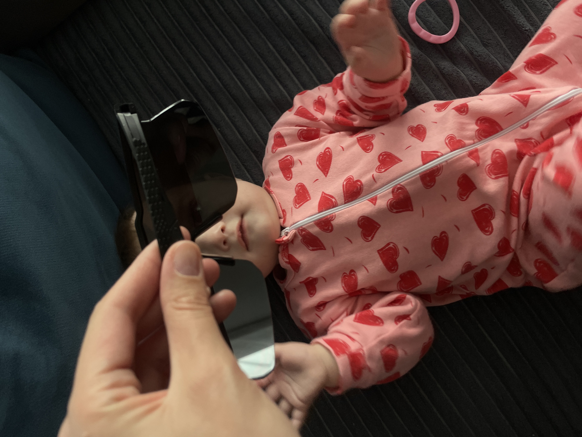 A baby, clad in a pink footed onesie adorned with numerous red heart patterns, lies on its back on a dark gray, ribbed surface. An adult's hand is prominently featured in the foreground, holding a pair of black sunglasses near the baby's face. The baby's head is slightly turned towards the sunglasses, with its eyes appearing closed or softly looking upward, and its mouth slightly open.
The pink onesie has a white zipper running down its front. One of the baby's feet is slightly raised, and its small hands are visible. A light pink, ring-shaped toy or teether rests on the dark gray surface near the baby's right side. The background also includes a section of dark blue fabric to the left.
The scene captures a tender, playful moment, likely indoors within a home setting, perhaps in Подшивалово, Russia. There is no visible text in the image.