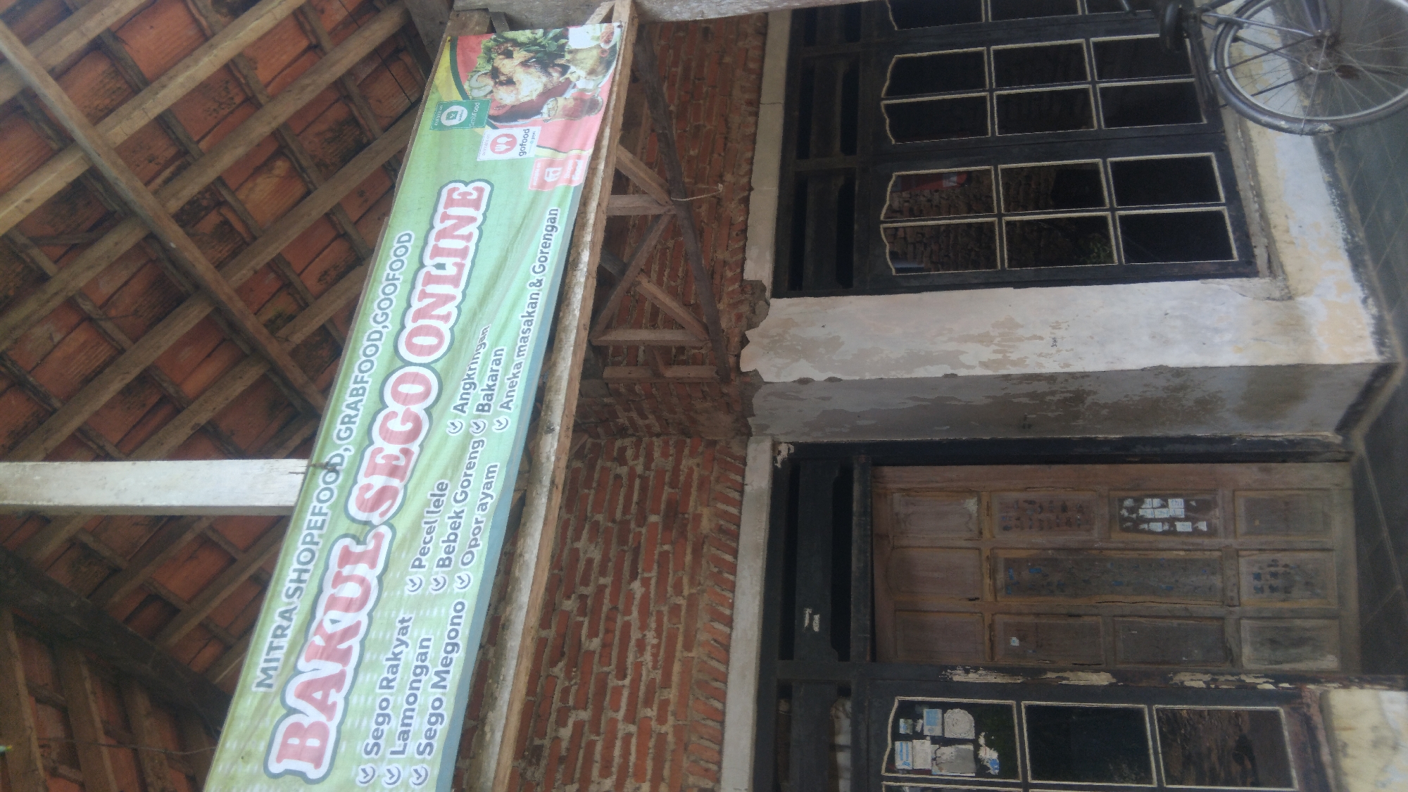 A vertical, eye-level, medium shot captures the facade of a food establishment in Watesalit, Indonesia. A green banner with white lettering is strung across the top of the building, advertising "MITRA SHOPEFOOD, GRABFOOD, GOOFOOD BAKUL SEGO ONLINE". Below the banner, the brick walls of the building are visible, with a wooden door and glass-paned windows on either side. The door is made of weathered wood and has multiple rectangular panels, some of which contain small printed advertisements. The windows are framed in dark wood, and the glass panes reflect the interior of the building. To the right of the windows, a portion of a bicycle wheel and frame is visible, suggesting it is parked nearby. The building appears to be old, with peeling paint on the walls and signs of wear and tear on the wooden elements. The roof structure, made of wooden beams and terracotta tiles, is visible above the banner. The overall scene depicts a humble, local eatery in a rural or semi-urban Indonesian setting.