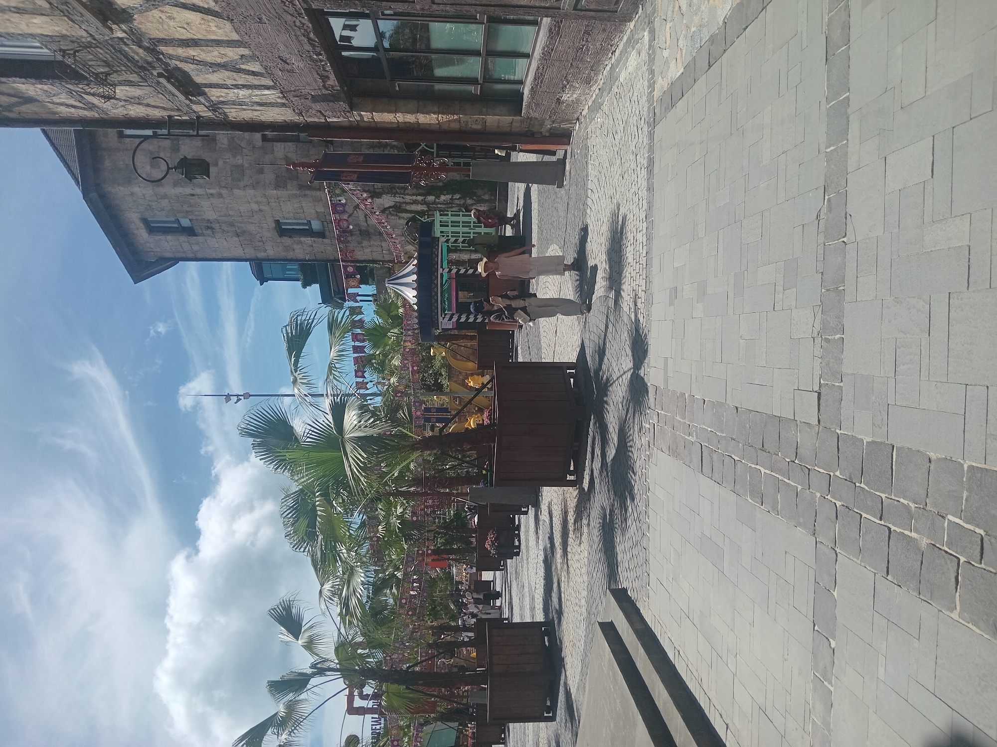 The image depicts an outdoor scene in a European-style village with cobblestone pathways and stone buildings. Palm trees are planted in large wooden planters, casting shadows on the ground. In the middle ground, two women are walking. One is wearing a hat and a long dress, while the other wears a dark jacket and light-colored pants. Banners and decorative flags are visible strung between buildings, suggesting a festive atmosphere. The architecture features stone facades with timber accents and ornate lampposts. The sky is blue with scattered clouds. The location is identified as a street in the Bà Nà Hills resort area in the city of Đà Nẵng, Vietnam, a popular tourist destination known for its French-inspired village.