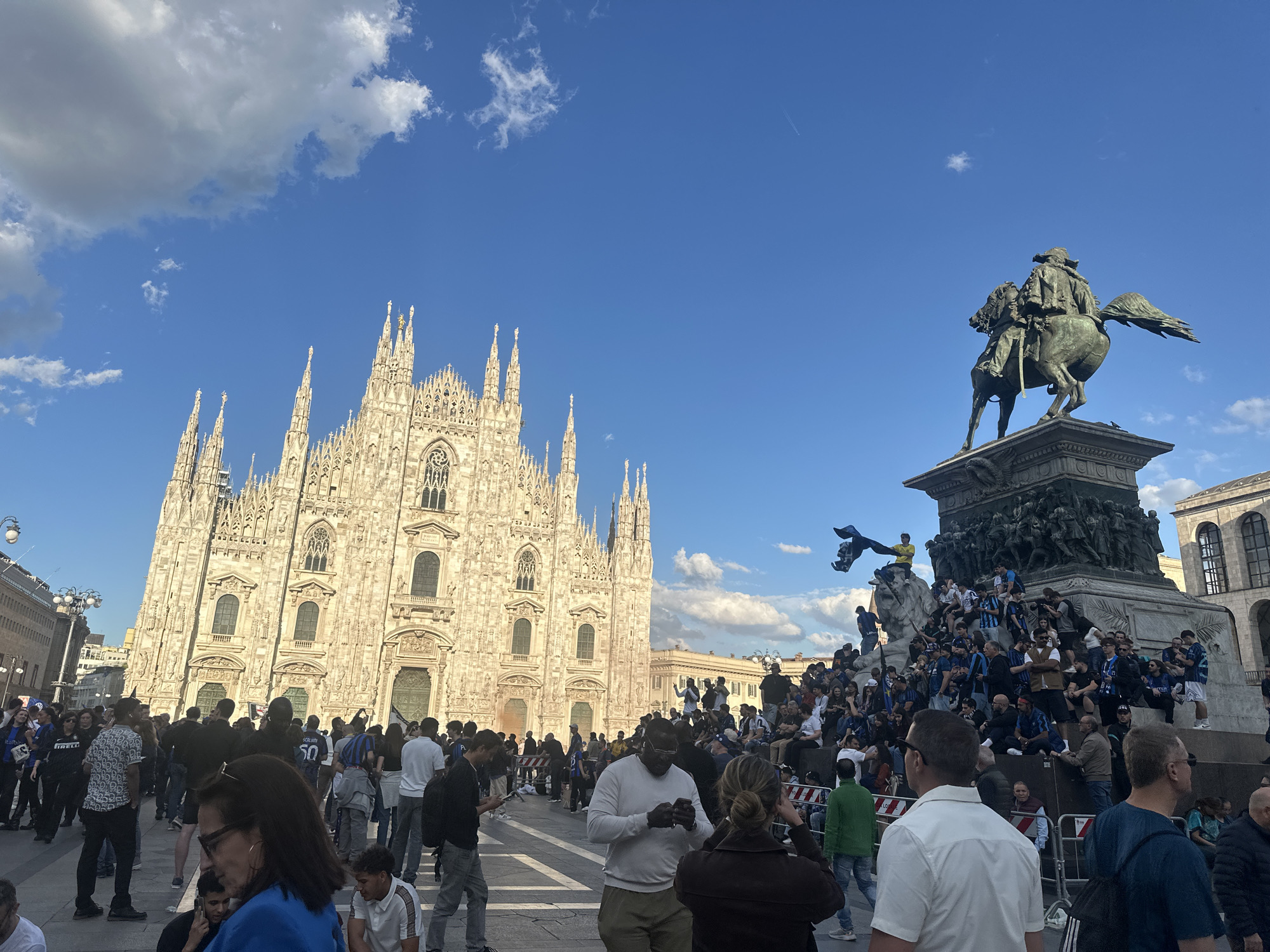 The image shows a large crowd of people in the Piazza del Duomo in Milan, Italy. In the background is the Duomo di Milano, the city's cathedral. To the right of the frame is the statue of Vittorio Emanuele II, the first king of Italy, on horseback. Many people are gathered around and on the statue, some wearing blue and black clothing, suggesting they are fans of the Inter Milan soccer team. People are standing, sitting, taking photos, and interacting with each other. The sky is blue with scattered clouds.