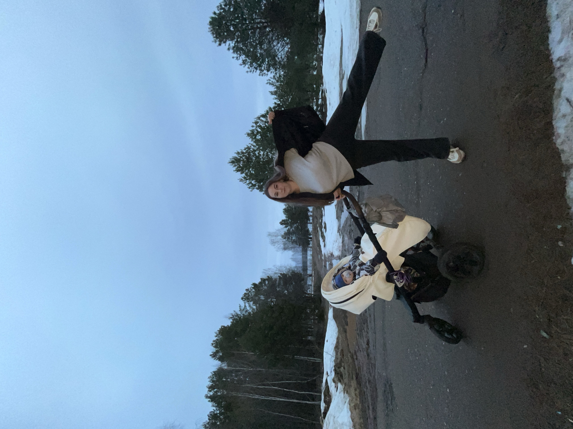 A young woman is seen posing outdoors with a baby stroller near the city of Chaikovsky, Russia. The woman is light-skinned with long brown hair, wearing a white shirt, black jacket, and black pants with white sneakers. She has one leg extended upward. The baby stroller is tan with a baby inside, wearing a blue hat. The environment appears to be a road or pathway bordered by patches of snow and trees. The sky is overcast.