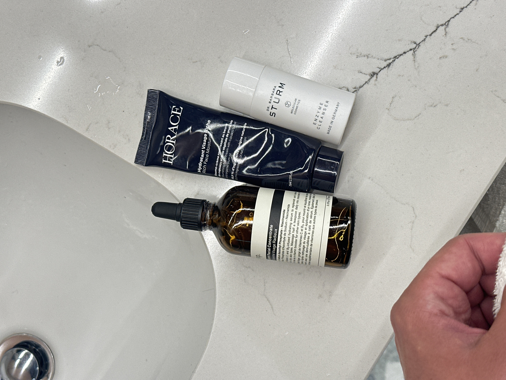 The image captures a close-up view of a bathroom countertop and sink area, likely in a residence in London, United Kingdom. Three different skincare products are neatly arranged on the light-colored, subtly veined countertop.
On the left, a white ceramic sink basin with a chrome drain is partially visible. Small water droplets are scattered on the countertop near the sink, suggesting recent use.
The products include:
1. A dark blue tube labeled "HORACE Hydratant Visage Riche Rich Face Moisturizer," with French and English text describing it as a "Baume-crème nourrissant au beurre de cupuaçu et huile végétale de prune bio" (Nourishing balm-cream with cupuaçu butter and organic plum oil), containing "99.9% d'ingredients d'origine naturelle" (99.9% of natural origin) and is "75ml 2.5fl.oz."
2. A white cylindrical bottle with a white cap, labeled "DR. BARBARA STURM MOLECULAR COSMETICS ENZYME CLEANSER MADE IN GERMANY."
3. An amber glass dropper bottle, partly obscured but showing text like "Facial Concentrate Concentré Visage Radieux," listing ingredients "Sodium Ascorbyl Phosphate · Niacinamide," and describing it as a "Vitamin C and B3-infused hydrating serum to replenish and balance oily skin types" in both English and French, indicated as "3.3fl.oz."
On the right side of the frame, a person's hand is visible, holding a white, textured towel, suggesting a recent or upcoming cleansing routine. The overall impression is one of a clean and organized personal care space.