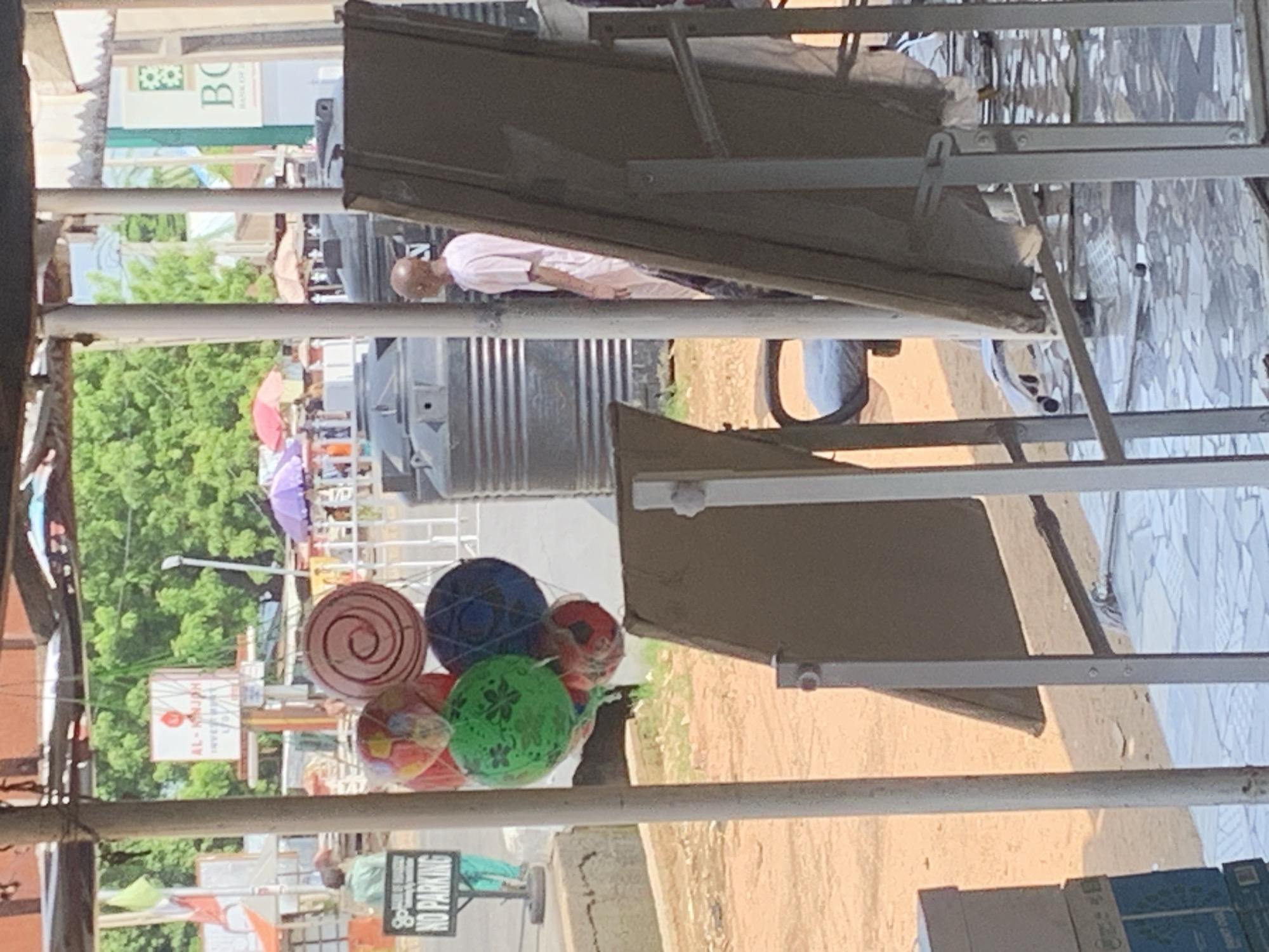 The image depicts an outdoor scene in what appears to be a market or a roadside stall area. In the foreground, a collection of colorful balloons are hanging, some are round and patterned, while one large swirled balloon is a prominent feature. To the left of the balloons, a "NO PARKING" sign is visible on a stand. A person wearing a green shirt and pants is walking in the background. Further back, large black water tanks are stacked. A bald man wearing a light pink garment is seen standing and facing away from the camera. In the background, there are buildings and lush green trees, suggesting a possibly warm climate. The ground appears to be a mix of dirt and paved areas. The overall impression is that of a bustling public space with various items for sale or display.