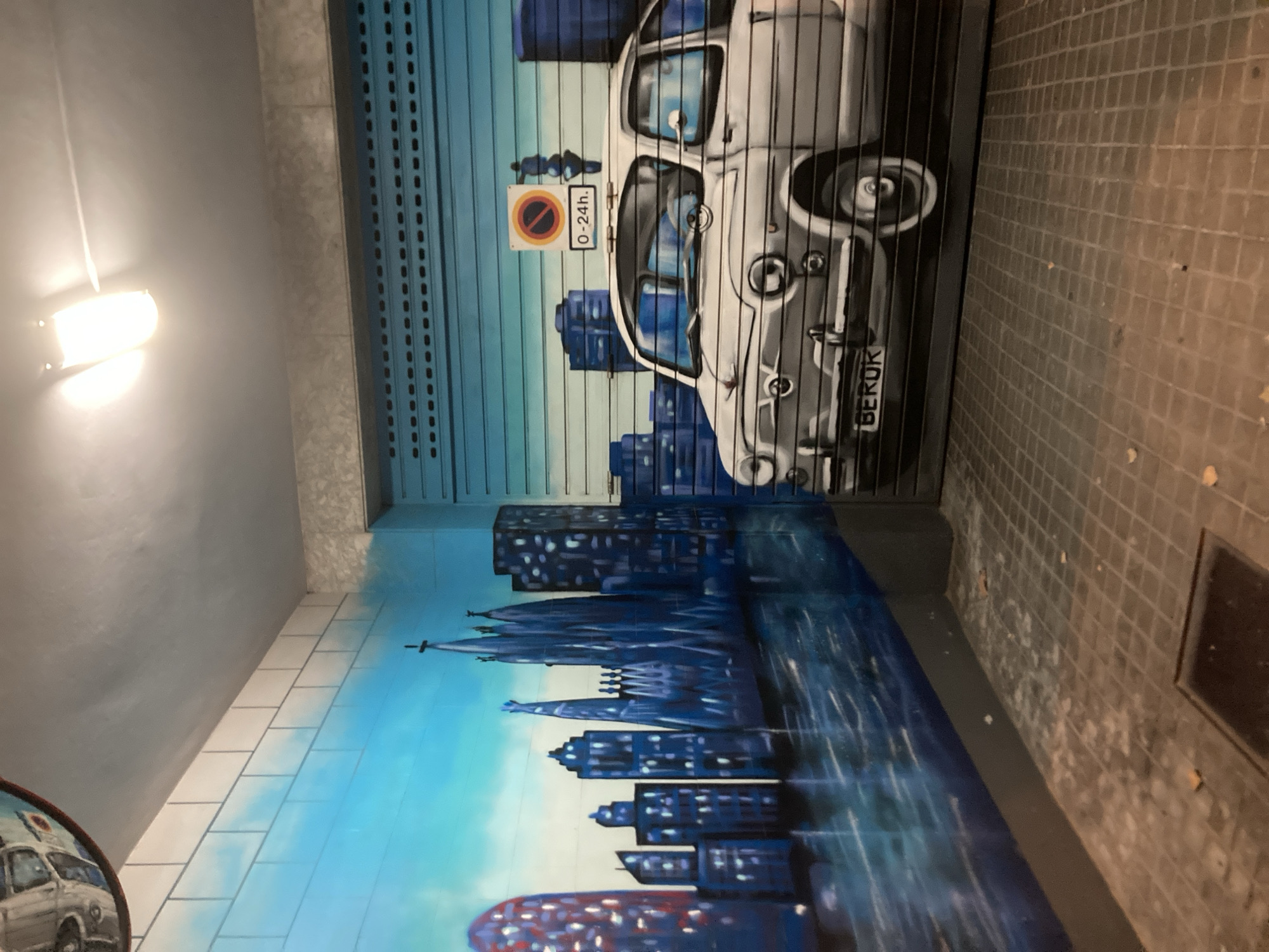 The image depicts a street art mural in Barcelona, Spain. The mural features a cityscape with iconic architecture, possibly representing the Sagrada Familia, on the left wall. The right wall is a roller shutter door painted with a vintage car, a sign indicating parking restrictions (0-24h), and a silhouette of a person. The overall scene is illuminated by artificial light sources, suggesting an indoor or covered outdoor space like a garage or an alleyway. The floor is tiled and appears to be well-worn.