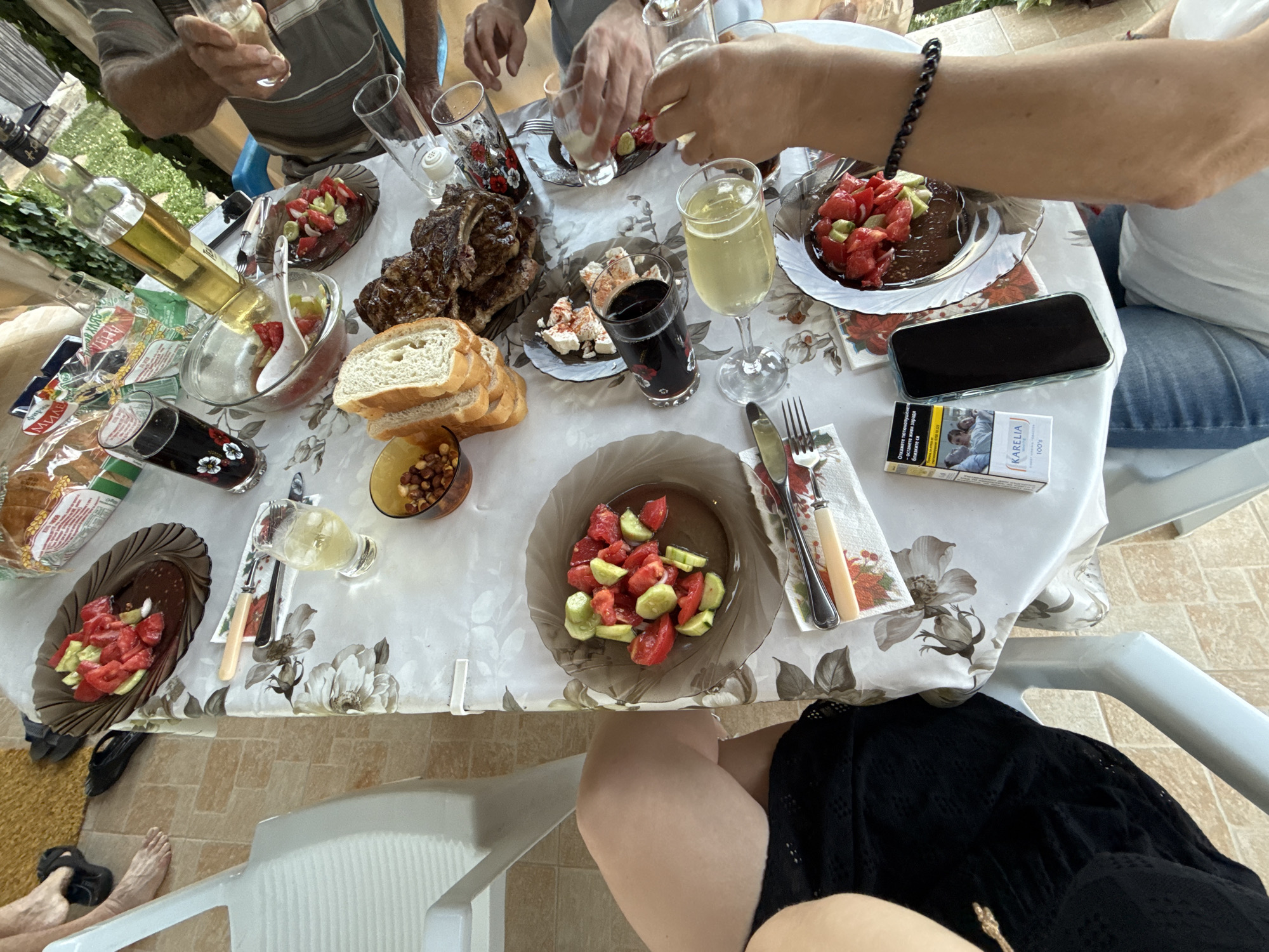 The image depicts an outdoor gathering at a table, possibly in Ravadinovo, Bulgaria, given the context provided. A table laden with food and drinks is the central focus. On the table, there is a large piece of grilled meat, sliced bread, a salad of tomatoes and cucumbers, a dish with what appears to be cheese and some red seasoning, nuts in a bowl, and various glasses filled with drinks, including what looks like lemonade and dark liquid. A bottle of olive oil stands upright. Two hands are visible actively pouring drinks into glasses, suggesting an ongoing toast or serving. In the foreground, a person wearing blue jeans and a white t-shirt is partially visible, with a mobile phone and a pack of Karelia cigarettes on the table nearby. The table is covered with a floral-patterned tablecloth. The background shows greenery and hints of an outdoor setting. The overall scene conveys a casual meal and social interaction.