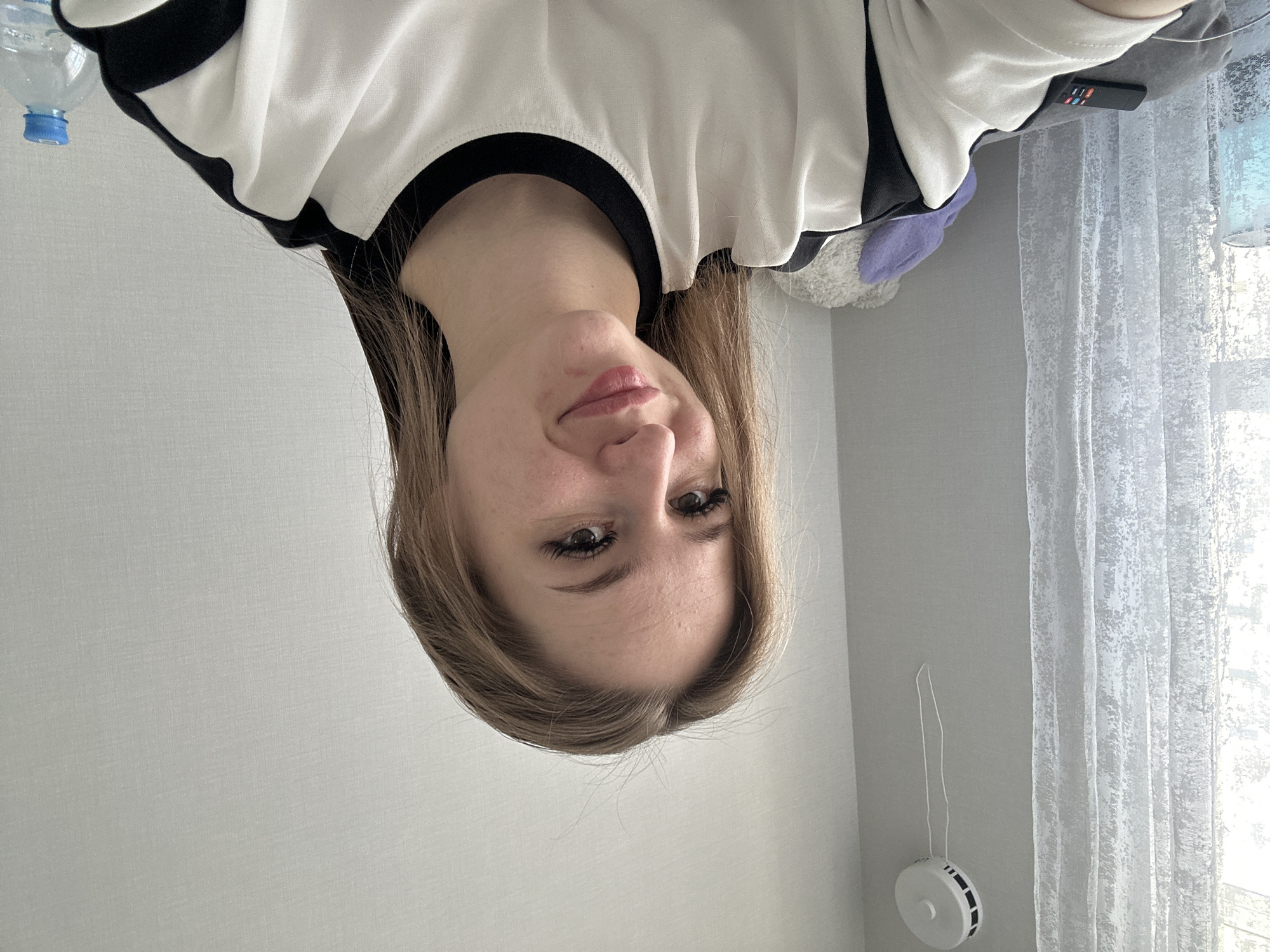 A young woman is depicted in a vertically inverted image, looking directly at the camera. Her head and upper torso are visible, with her light brown hair falling around her shoulders. She has dark eyes enhanced with eyeliner and mascara, and her lips are a soft pink with a slight upward curve, suggesting a neutral or gently smiling expression. She is wearing a white garment with a black collar and black trim on the shoulders.
The background consists primarily of plain, light grey walls. In the upper left corner, a clear plastic water bottle with a blue cap is partially visible. On the right side of the image, a sheer white curtain with a textured pattern covers a window, allowing natural light into the room, indicating it is daytime. A black remote control with colorful buttons rests on a grey surface near the top right. Further down the right wall, a white, round device, possibly a smoke detector or sensor, is attached to the wall, with a thin white cord hanging from it. Portions of what appear to be soft, plush items, one purple and one grey/white, are faintly visible behind her right shoulder. No explicit text is visible in the image.
