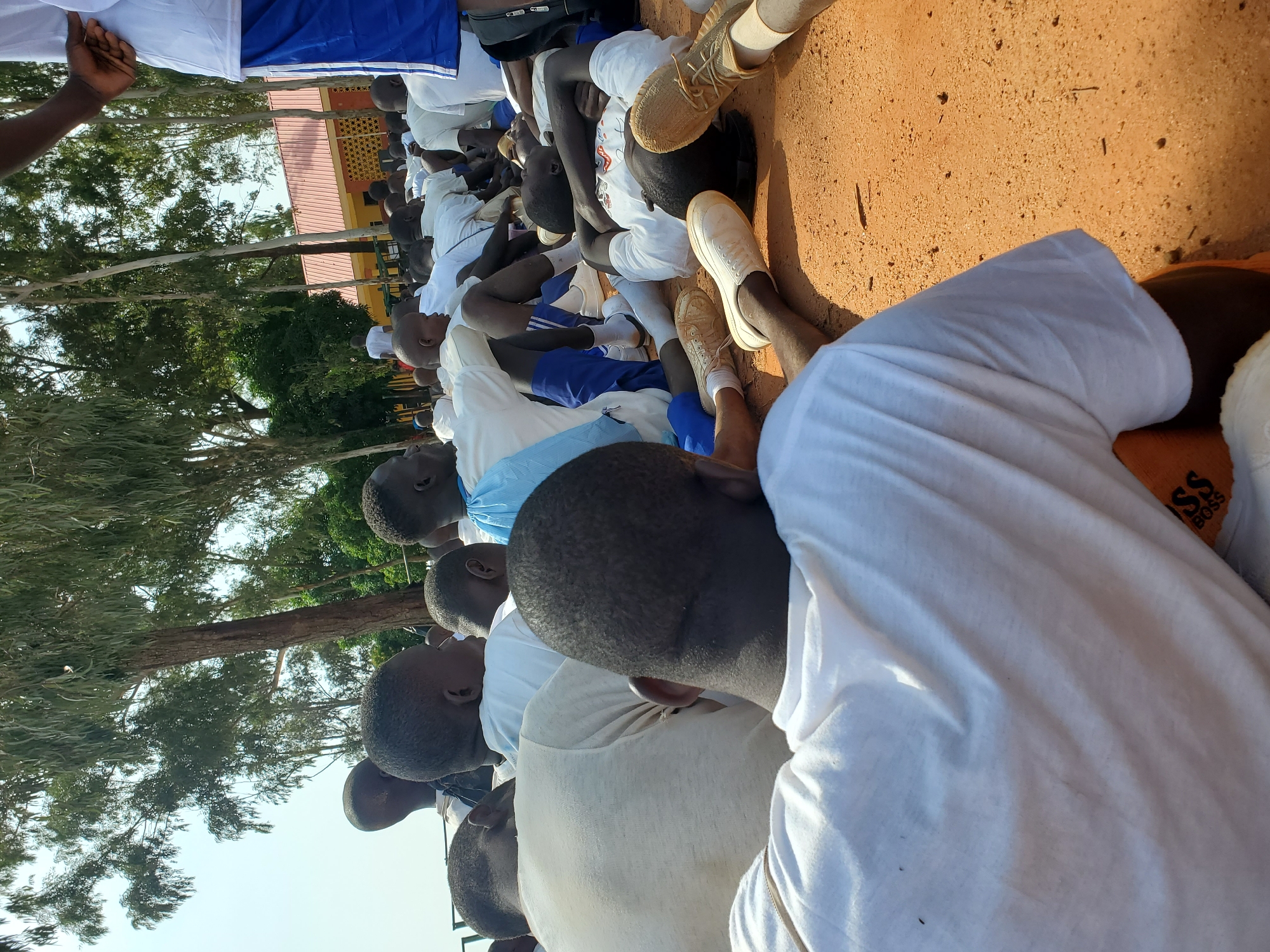 In Barakin Rafin Gora, Nigeria, a group of young people, predominantly boys and young men, are seated on the ground under trees. Many are wearing white t-shirts and some are in blue shorts. The ground is covered in a reddish-brown dirt. In the background, there is a building with an orange roof and a fence. The overall scene suggests an outdoor gathering or activity, possibly related to a school or training program.