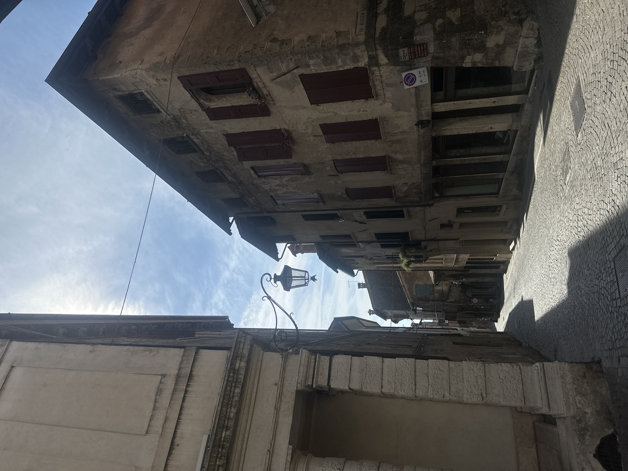 This is a vertical, eye-level, full shot of a cobblestone street in Maser, Italy, under a partly cloudy sky. The left side of the image is dominated by the corner of a light-colored stone building with large, arched openings. The right side features an aged, multi-story building with stucco walls, dark wooden shutters, and arched windows. A wrought-iron street lamp hangs from a bracket attached to the stone building on the left, extending over the street. The street is paved with small, gray cobblestones that reflect the sunlight. Shadows from the buildings stretch across the road. In the distance, the street curves and narrows, lined with more buildings. A street sign is visible on the right-hand building, with Italian text and a parking symbol. The overall atmosphere is one of historic European architecture bathed in sunlight.
