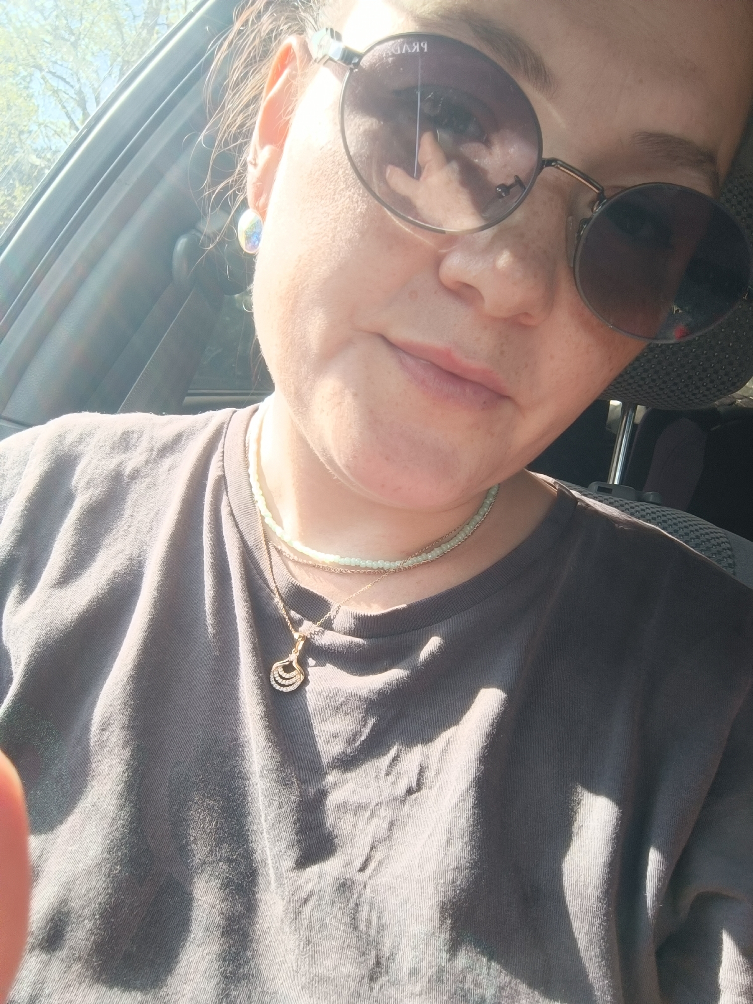 A close-up, waist-up selfie of a woman in a car is captured, with the right side of her face and upper body filling the frame. She is wearing round, dark-lensed sunglasses, and her hair is visible on the left side of her head, partially obscuring her ear. A small, iridescent stud earring is visible in her right ear. The woman has fair skin with freckles across her nose and cheeks. She is wearing a plain, dark grey t-shirt, and two necklaces adorn her neck. The top necklace is a thin, light-colored beaded chain, and the bottom one is a gold chain with a delicate pendant. Sunlight streams in from the left, creating bright highlights and shadows across her face and shirt. The background shows the interior of a car, including the window, car door, and a headrest. The environment suggests a sunny day. There is no information available to determine the specific location within Foki, Russia.