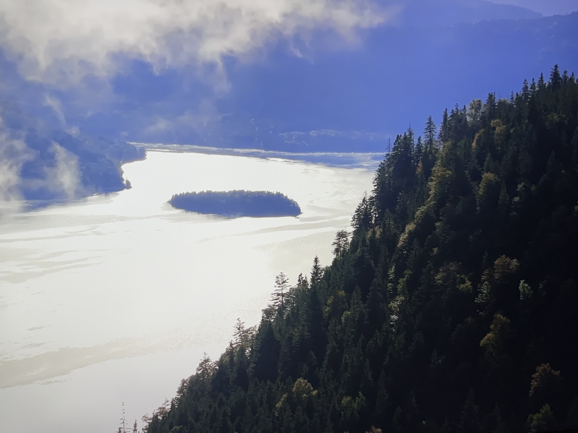 The image depicts a vast body of water, possibly a lake or sea, reflecting the bright sky and scattered clouds. In the distance, a small, tree-covered island is visible. To the right of the water, a densely forested mountainside slopes down towards the water's edge. The trees are predominantly evergreen, with some scattered deciduous trees showing hints of autumn colors. The sky above is a vibrant blue, partially obscured by soft, white clouds that cast a diffused light on the scene. The overall impression is one of natural, serene beauty. The image does not depict the city of Burgas, Bulgaria.