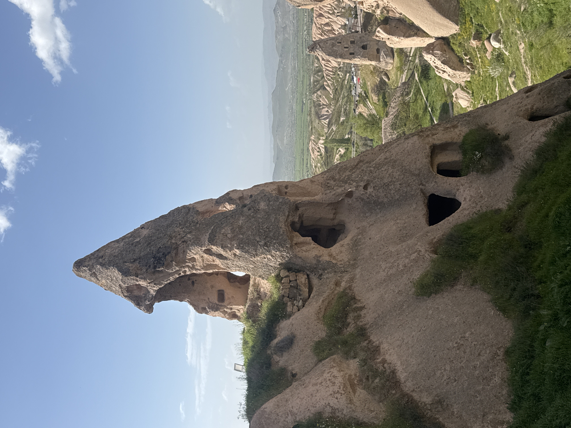 This is a landscape photo taken in Uçhisar Beldesi, Turkey. The foreground is dominated by a large rock formation with several caves carved into it. Some of these caves appear to be artificially made, while others might be natural erosion. Patches of green vegetation grow on the rock and at its base.
In the background, a valley unfolds with agricultural fields and other rock formations similar to the one in the foreground. The valley is a mix of green fields and rocky terrain. Further in the distance, the horizon is marked by a haze, suggesting the presence of distant mountains.
The sky is mostly blue with some scattered clouds. The overall light suggests it is daytime, with the rock formation casting shadows.
