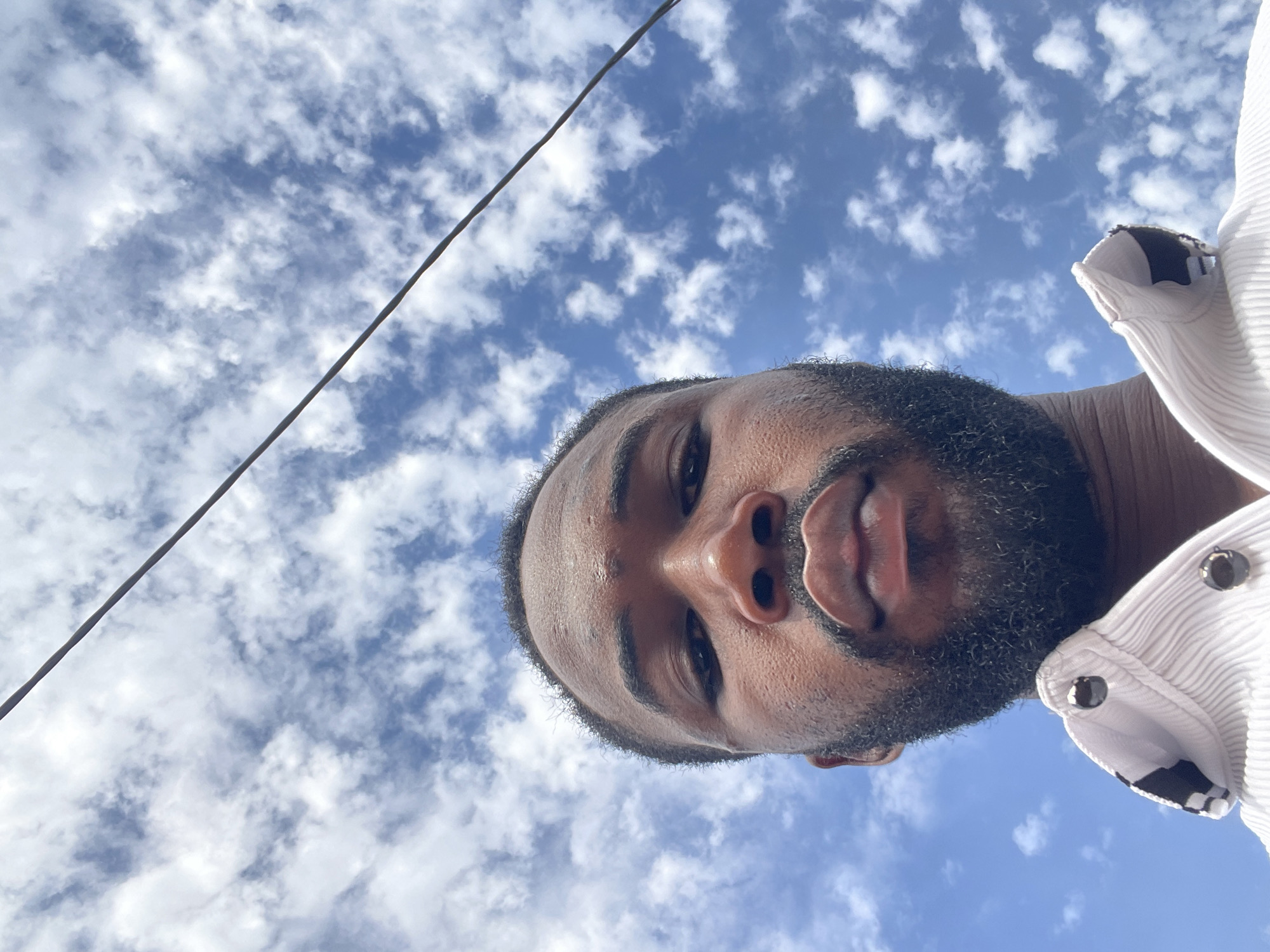 A low-angle selfie of a man with dark skin, a beard, and short dark hair, wearing a light-colored shirt with buttons on the collar. The background is a blue sky with scattered white clouds. A power line stretches across the upper left portion of the image. The location cannot be determined as Lokoja, Nigeria based only on the visual information in the image.