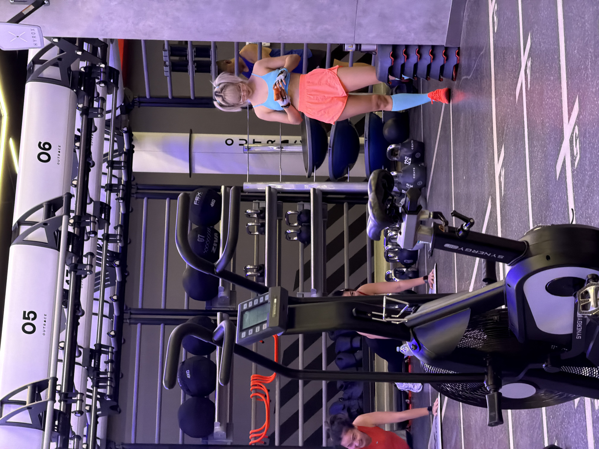 The image depicts the interior of a modern gym, likely located in Nong Pa Khrang, Thailand, given the provided context. In the foreground, a black exercise bike with the "SYNERGY" logo is visible. To the left, a woman wearing a red tank top and dark leggings is in a plank position on a mat. To the right, a woman with blonde hair styled in pigtails, wearing a blue sports bra, coral shorts, and light blue compression socks, is standing and looking at her orange mobile phone. She is also wearing glasses and bright orange athletic shoes. Behind her, a stack of black weight plates with red accents is present. In the background, shelves are laden with various gym equipment, including black medicine balls and kettlebells. Overhead, a structure with numbered stations ("05" and "06") is visible, suggestive of a fitness class or training area. The overall environment is brightly lit with artificial lighting, and the floor is marked with white lines.