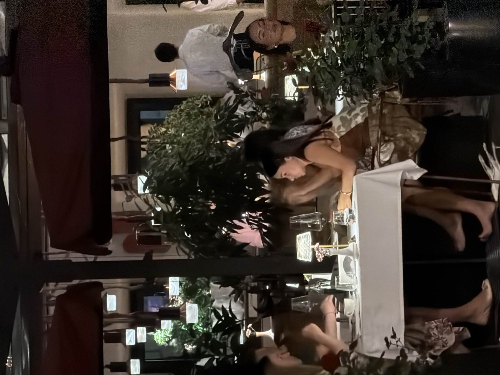 The image depicts an interior scene, possibly a restaurant in Moscow, Russia, based on the architectural style and ambiance. Multiple individuals are present, engaged in dining. The foreground features a table setting with glasses, plates, and a white tablecloth. Several people are seated around the table, including a woman with dark hair, possibly interacting with a mobile phone. Other individuals are visible in the background. Artificial lighting is evident, creating a warm atmosphere. Plants are interspersed throughout the space, adding a touch of nature to the indoor setting.