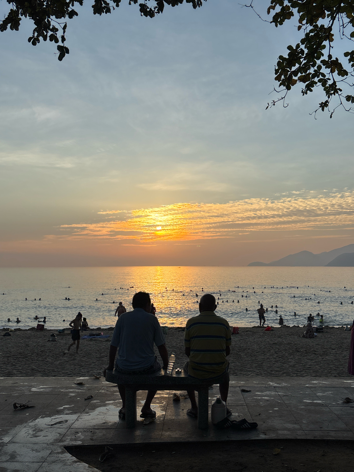 This is a full-frame, eye-level shot of a beach scene at sunset in Nha Trang, Vietnam. In the foreground, two men sit on a concrete bench facing away from the camera, looking out at the ocean. One man wears a blue t-shirt and shorts, while the other wears a yellow and blue striped t-shirt and shorts. There are some discarded items, including a bottle of water and a pair of flip-flops, on the ground near the bench.
The middle ground shows a sandy beach with numerous people swimming and wading in the ocean. The water reflects the warm hues of the setting sun. In the background, the sun is low on the horizon, casting a golden glow across the sky and water. The sky is partly cloudy with soft, wispy clouds. Mountains are visible in the far distance. Overhanging branches with leaves frame the top of the image, creating a natural border. The overall impression is one of a peaceful evening by the sea.