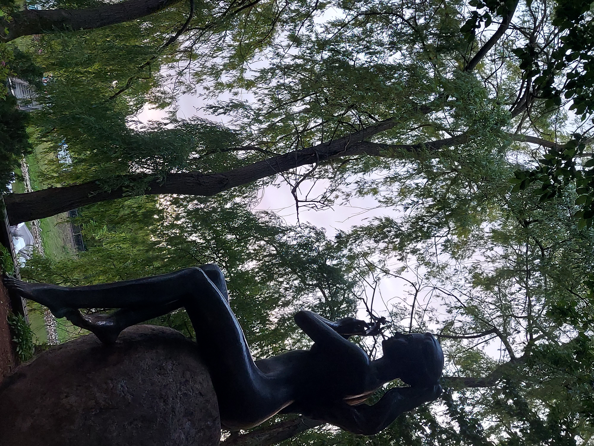 The image shows a bronze sculpture of a nude woman sitting on a large rock, set amidst weeping willow trees. The sculpture is dark and silhouetted against a light sky, with its profile facing right. The woman has her hair pulled back in a ponytail and is holding something to her face, possibly a flower or a leaf. Her legs are crossed, and her feet are visible. The weeping willows create a dense green canopy, with their long branches hanging down. In the background, a body of water can be seen, along with some man-made structures, including what appears to be a wooden bridge or walkway. This sculpture is located in Brest, Belarus, and is situated in a park-like setting. The overall scene suggests a serene and natural environment.