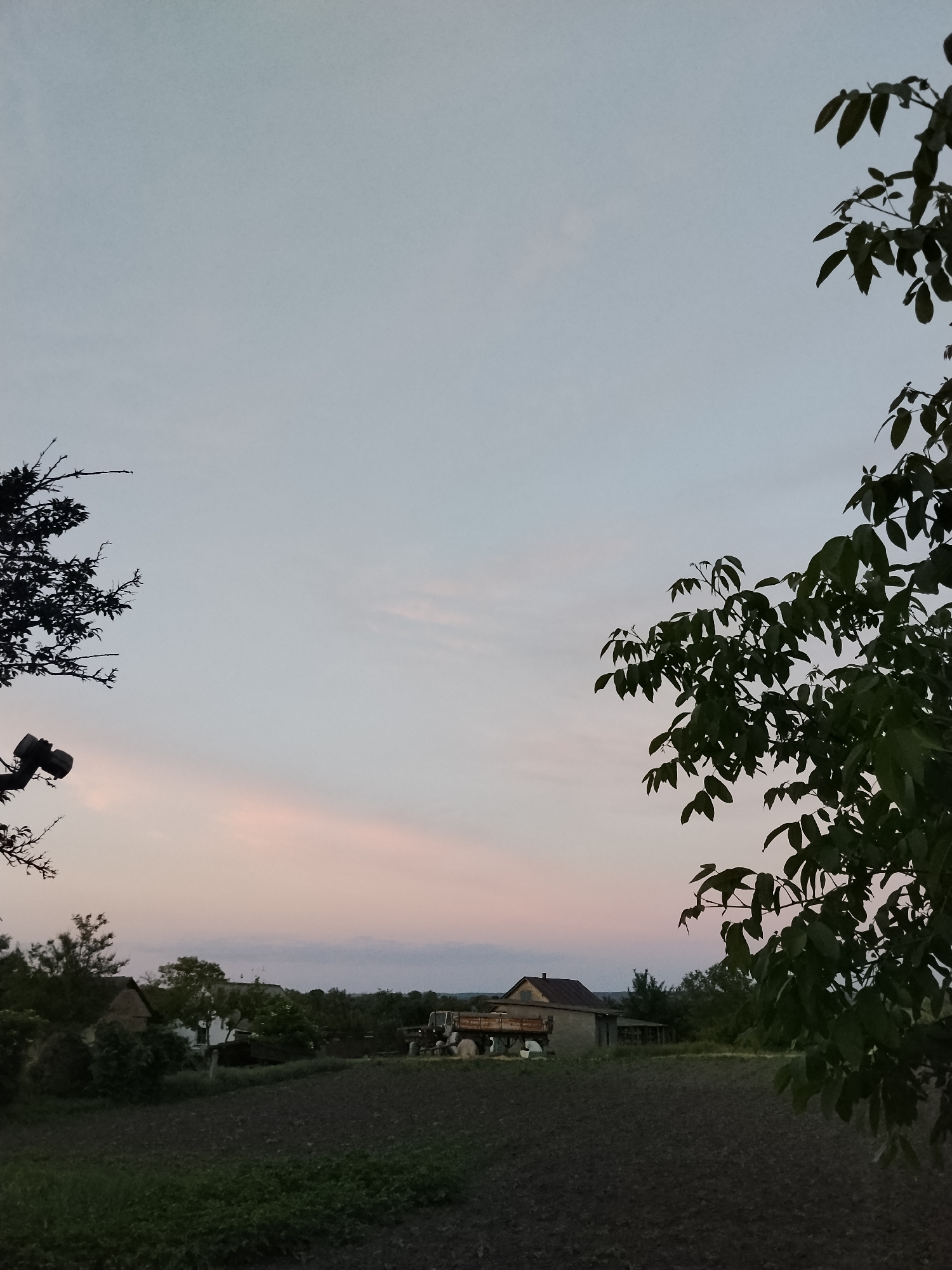 This media file captures a serene rural landscape in Ustyvytsia, Ukraine, likely at dusk or dawn. The sky dominates the upper portion of the image, displaying a soft gradient from a light blue-grey at the top to delicate pale pink and orange hues closer to the horizon, indicating the fading or rising sun. A few wispy clouds are subtly visible against the sky.
The scene is framed by dark silhouettes of leafy tree branches in the left and right foreground. On the left, partially obscured by the branches, is a dark object resembling a security camera or floodlight mounted on a pole or trunk.
In the middle ground, a tilled field or garden plot stretches across the width of the image, showing dark, rich earth with sparse green vegetation. Beyond the field, several buildings are visible. A prominent structure, possibly a house or farm building, features a dark brown roof and what appears to be a balcony or raised porch area. A white satellite dish is attached to one of the buildings. Further into the background, other smaller structures and dense tree lines complete the rural setting.
No people are visible in the image, and there are no explicit activities taking place, contributing to the peaceful and quiet atmosphere. No visible text is present. The overall impression is one of tranquility in a quiet countryside setting as day transitions to night or vice versa.