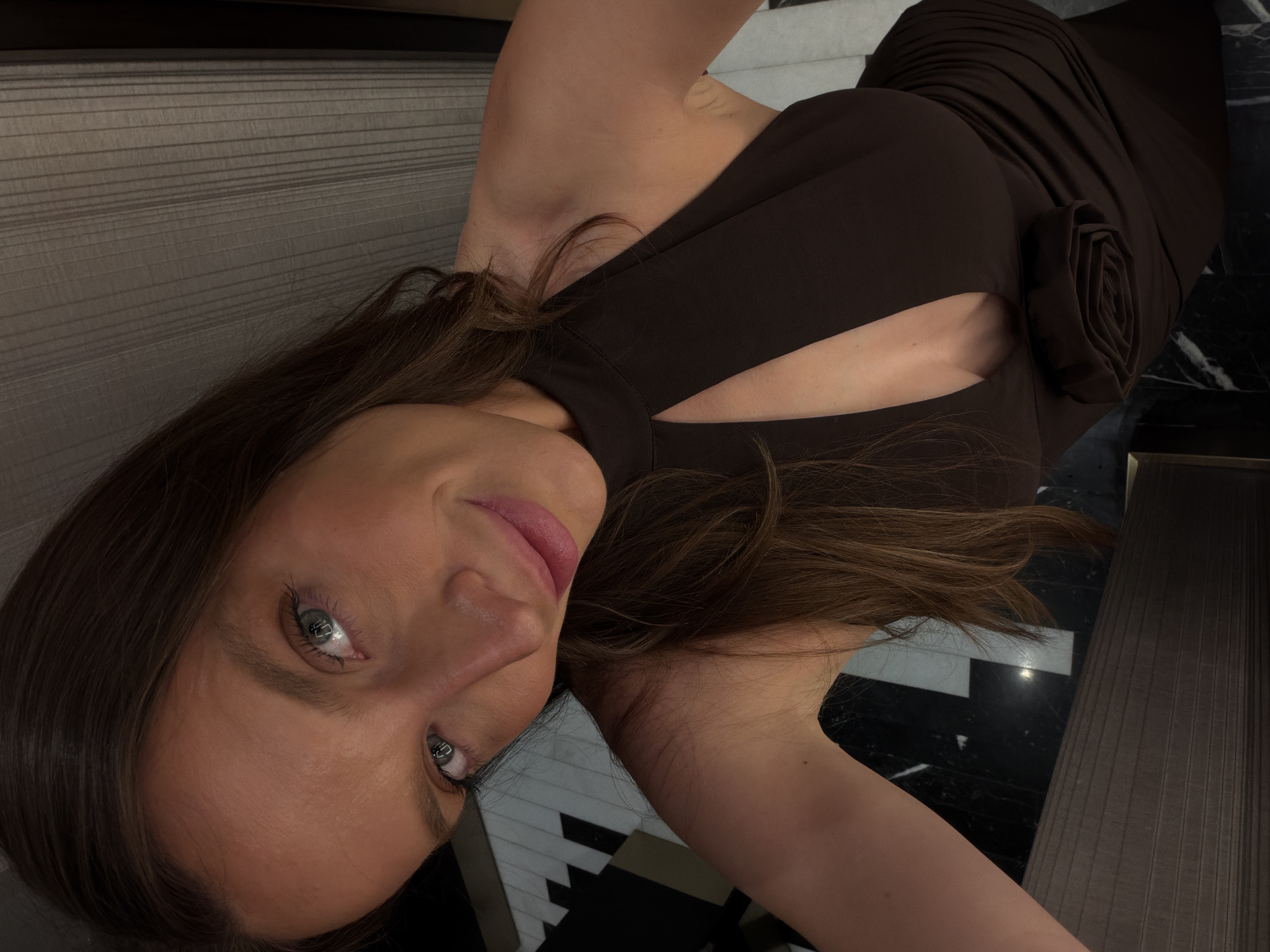 A female individual is depicted in a close-up, upward-angled portrait, likely a selfie, within an indoor setting. She has long, dark brown hair, styled with a center part and falling over her shoulders. Her eyes are blue-grey, exhibiting distinct reflections of light sources. Her lips are a soft pink-mauve, curved in a subtle smile. She is wearing a dark brown, possibly black, halter-neck dress featuring a keyhole cutout below the collarbone and a sculptural fabric rose appliqué on the left side of her torso.
The background consists of an interior space with ambient, somewhat dim lighting. To the viewer's left, behind her, a section of floor or wall is visible, patterned with alternating black and white geometric shapes, indicative of marble or tilework. To the viewer's right and behind her, a textured surface, possibly a wall panel or curtain, displays fine vertical lines in a dark grey-brown tone. The floor in the lower sections of the image is dark and reflective, appearing to be polished stone or marble. No specific landmarks or unique architectural elements are visible to identify the location as Budva, Montenegro.