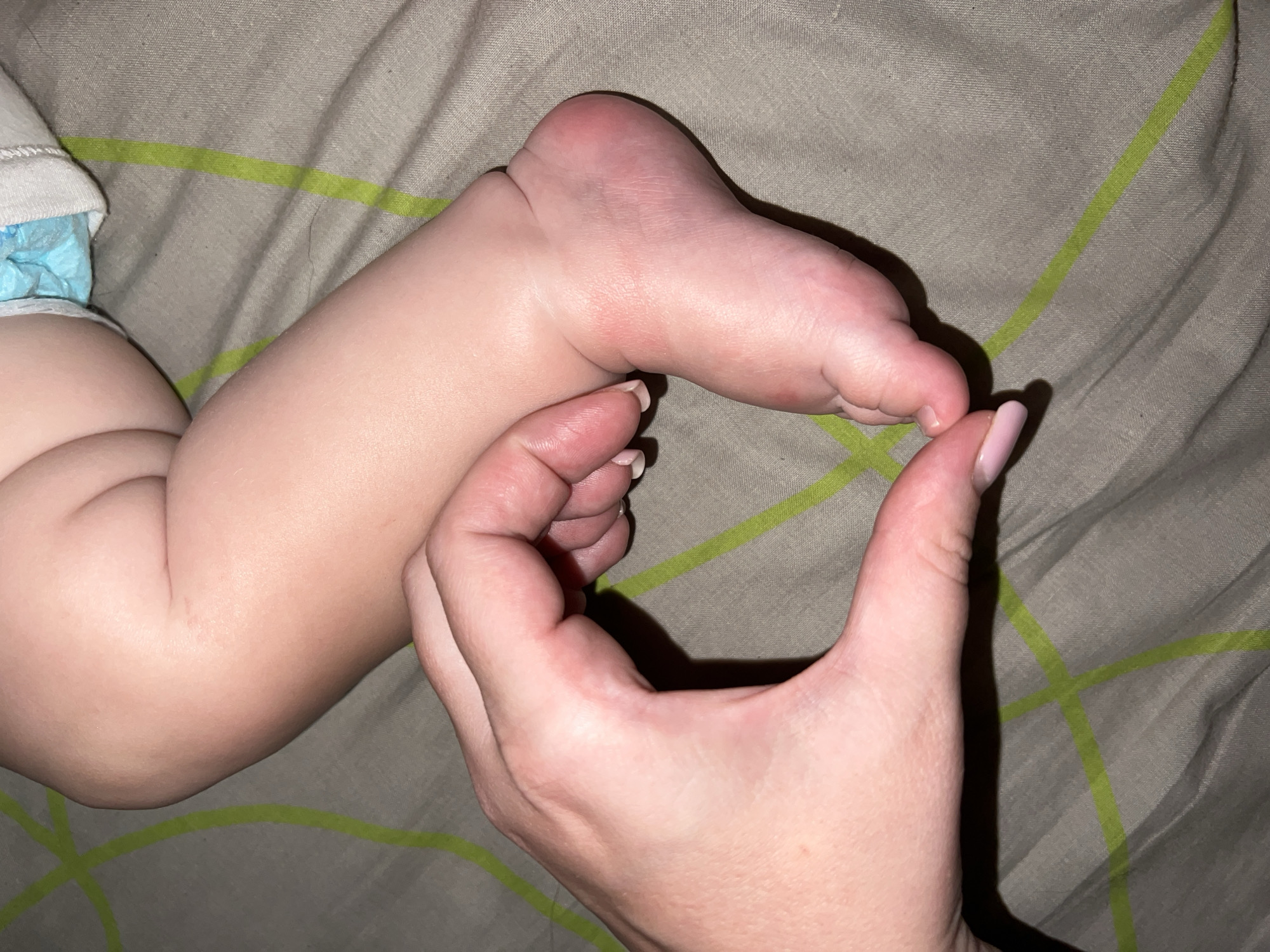 A close-up, overhead shot shows an adult's hand forming a heart shape around a baby's foot and lower leg. The baby's skin is pale and smooth, with dimples on the thigh and ankle. A blue diaper is visible at the top of the frame. The background is a brown fabric surface with a green geometric pattern. The scene is static and peaceful.