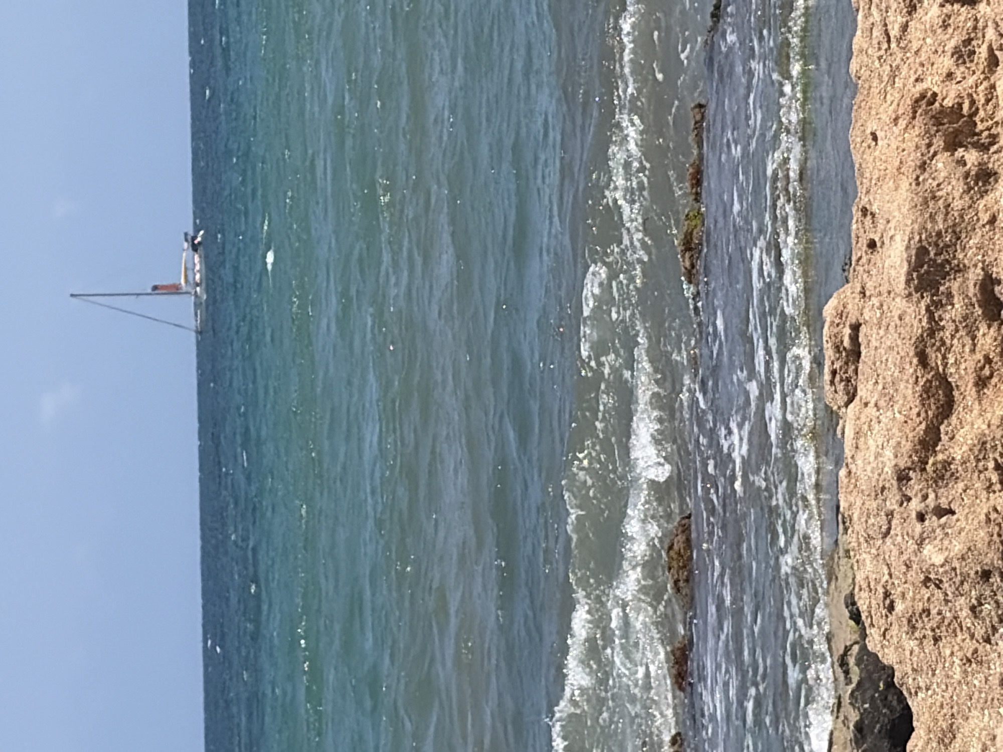 This media file shows a close-up view of the sea and a rocky shore. The water is a mix of blue and green, with gentle waves lapping at the shore. The shore itself is made up of large, rough-textured rocks in shades of brown and tan. A small sailboat is visible in the distance on the left side of the frame, with a person on board. The sky above is a clear, light blue, suggesting it is daytime and the weather is fair. The overall scene evokes a sense of tranquility and natural beauty. No text is visible in the media file.