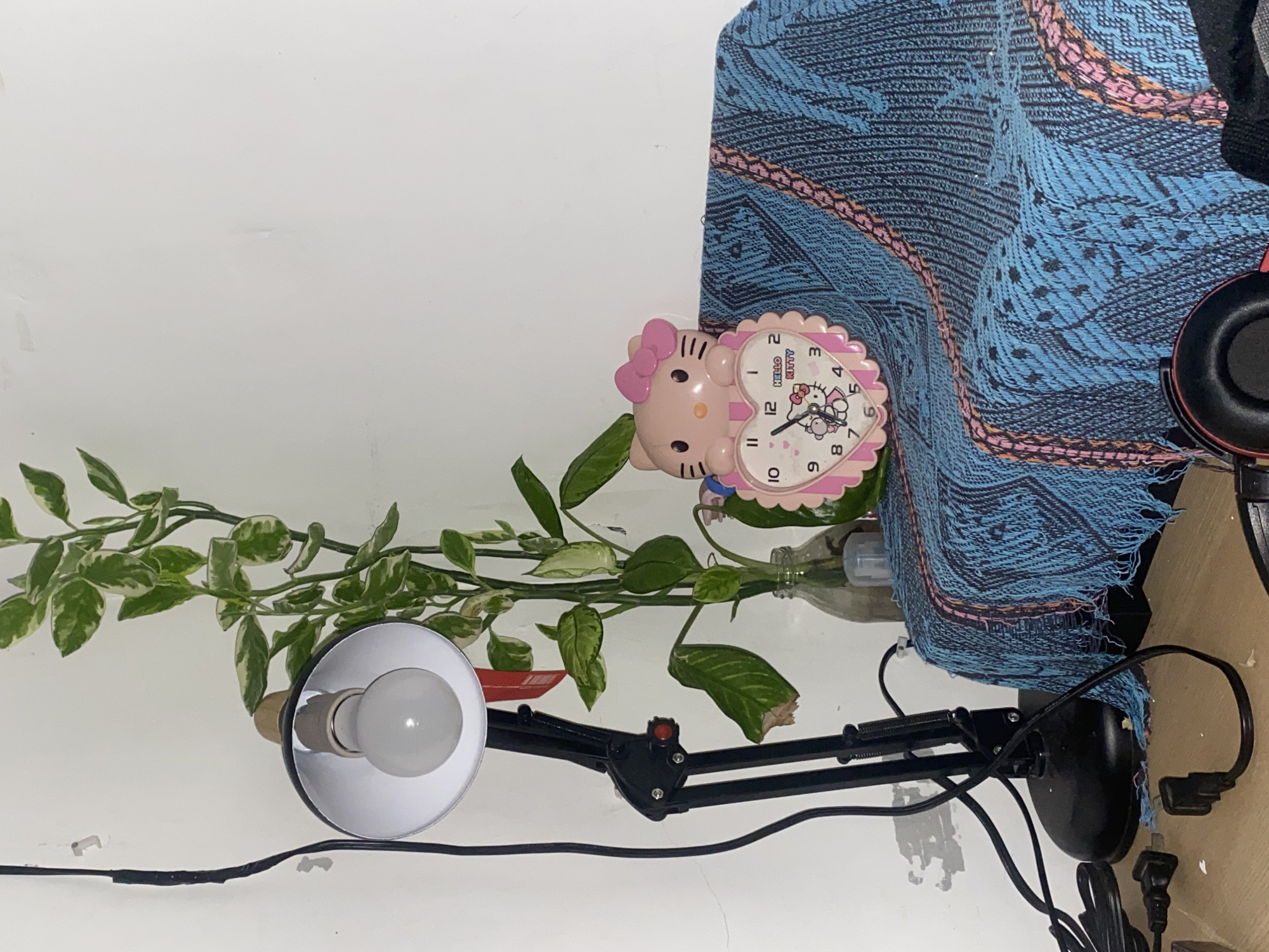 The image shows a close-up indoor scene. In the center, a pink heart-shaped Hello Kitty alarm clock rests on a blue and pink patterned fabric draped over an unseen object. To the left, a black adjustable desk lamp with a white bulb is positioned next to a tall green plant with variegated leaves in a clear glass vase. The background is a plain white wall with some scuff marks. The foreground shows a wooden surface with black electrical cords and plugs. This scene is likely a desk or bedside table setup in an urban setting.