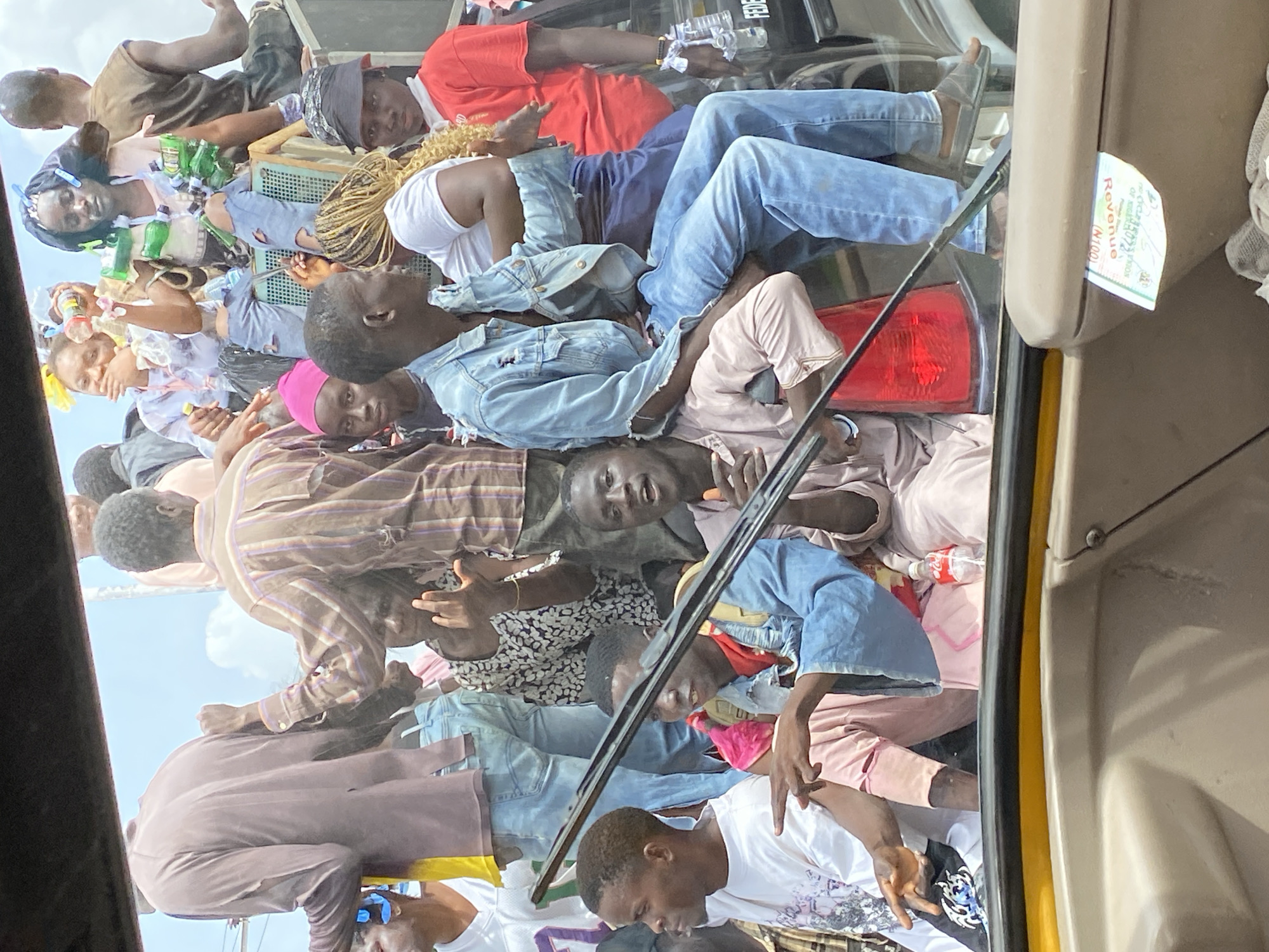 The image depicts a crowded vehicle, likely a taxi or bus, in Bukuru, Nigeria. Numerous people are piled on top of each other on the vehicle's roof. They are dressed in a variety of casual clothing. A windshield wiper is visible. The sky is partly cloudy. There's a "Revenue" sticker displayed in the car. Several drink bottles are visible among the people and cargo on top of the car.