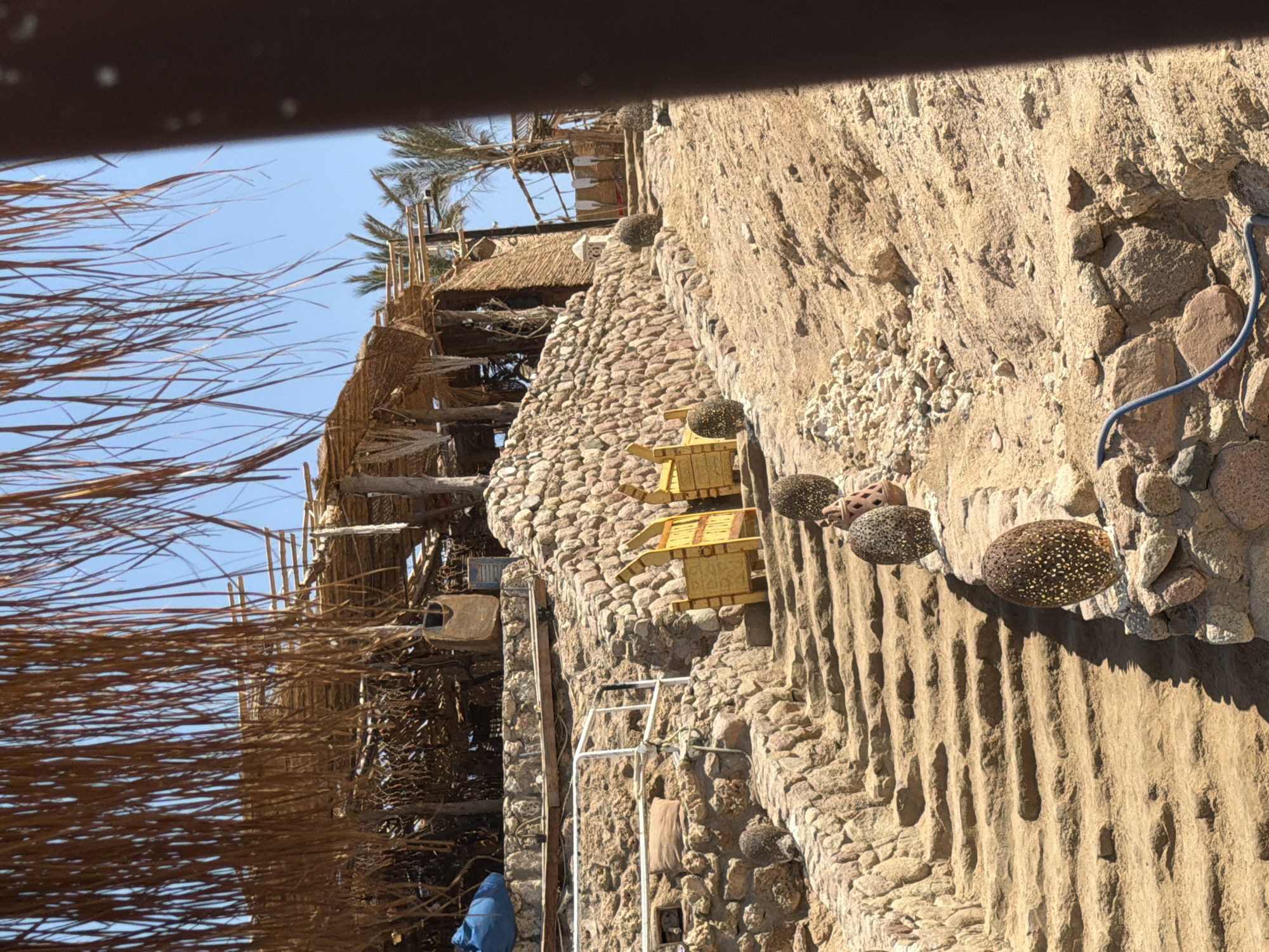 The image depicts a rustic outdoor restaurant or cafe situated on a sandy hillside in Sharm El Sheikh, Egypt. The structure is constructed primarily from natural materials, featuring a thatched roof supported by wooden poles, with walls made of woven reeds and rough stone. Several steps, also made of stone and packed sand, lead up the incline. Decorative, egg-shaped sculptures, some with intricate patterns and speckled surfaces, are placed along the side of the steps. Yellow wooden gates or partitions are visible near the mid-section of the stairs. Palm trees and other vegetation are interspersed around the structures. The overall impression is one of a relaxed, natural setting, possibly a beachside establishment.