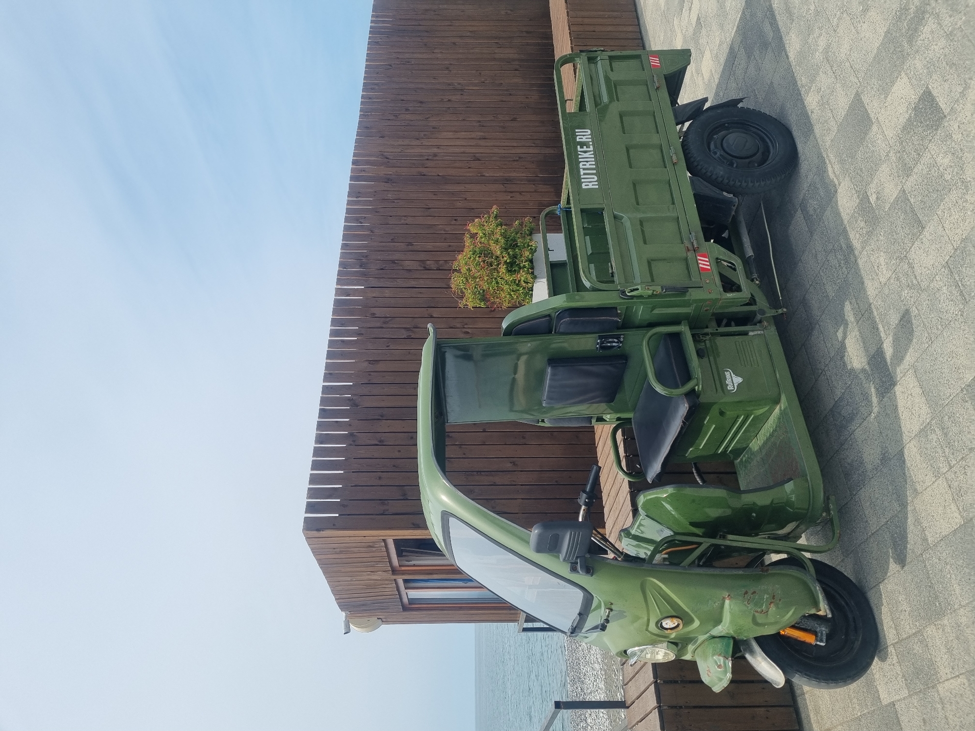 A green three-wheeled cargo vehicle, likely a mototrike or "rutrike," is parked on a paved surface in an outdoor setting. The vehicle features a driver's seat and a large cargo bed at the rear. The brand name "RUTRIKE.RU" is clearly visible in white lettering on the side of the cargo bed, accompanied by red and white striped warning stickers. The front of the vehicle shows minor signs of wear, including rust and small dents on the fender.
In the background, a modern building with dark wooden slat siding stands under a clear, bright blue sky. A vibrant bush with green and reddish leaves grows near the wooden structure. To the right, a body of water, consistent with the Black Sea in Sochi, Russia, is visible. The ground is paved with light-colored, patterned tiles. The scene is illuminated by bright daylight, suggesting a sunny day. No people are present.