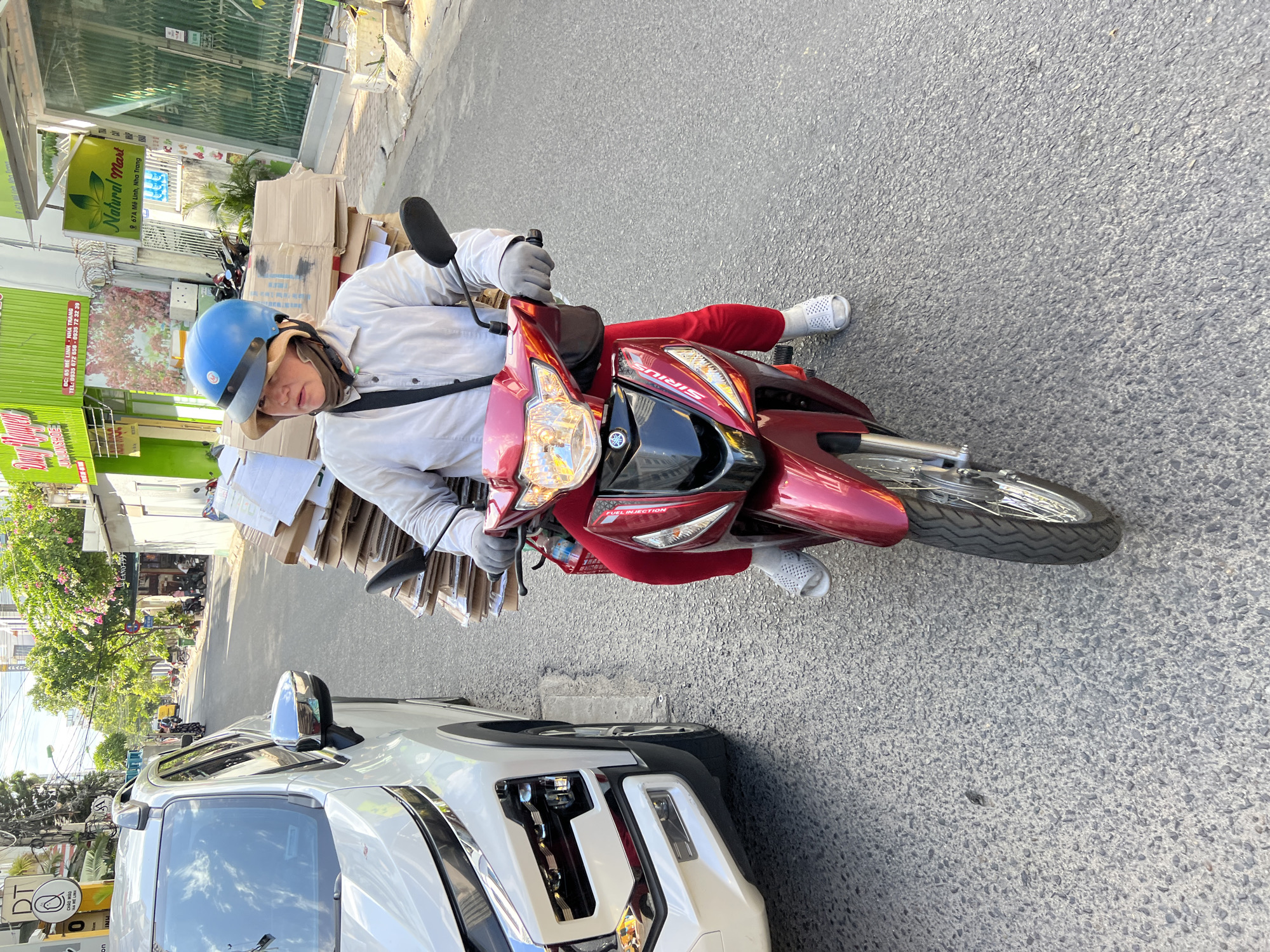 A woman wearing a blue helmet and a tan scarf is riding a red motorcycle loaded with cardboard boxes on a street in Nha Trang, Vietnam. She is wearing a light-colored long-sleeved shirt and red pants, with white slides on her feet. The motorcycle's front is visible, showing the Yamaha logo and the word "SIRIUS." To the left of the motorcycle is a white SUV. The background shows buildings, trees, and other motorcycles parked along the street, indicating an urban environment. A sign for "Natural Mart" is visible on a building in the background. The street is paved with asphalt.