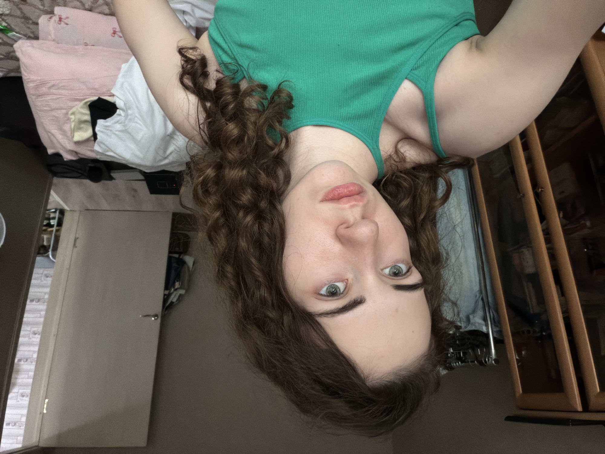 The image shows a young woman with curly brown hair and light-colored eyes, wearing a green tank top. She is positioned in what appears to be a bedroom. In the background, a wooden cabinet or wardrobe is visible to the left, and a doorway with a partially opened white door is on the right. The room contains clothing and bedding, suggesting a lived-in space. The lighting is dim, indicating it might be indoors with artificial light or natural light from a window. The image does not provide any specific clues or visual information that would allow for its location to be determined within the city of Tyumen, Russia, or any other specific geographical location.