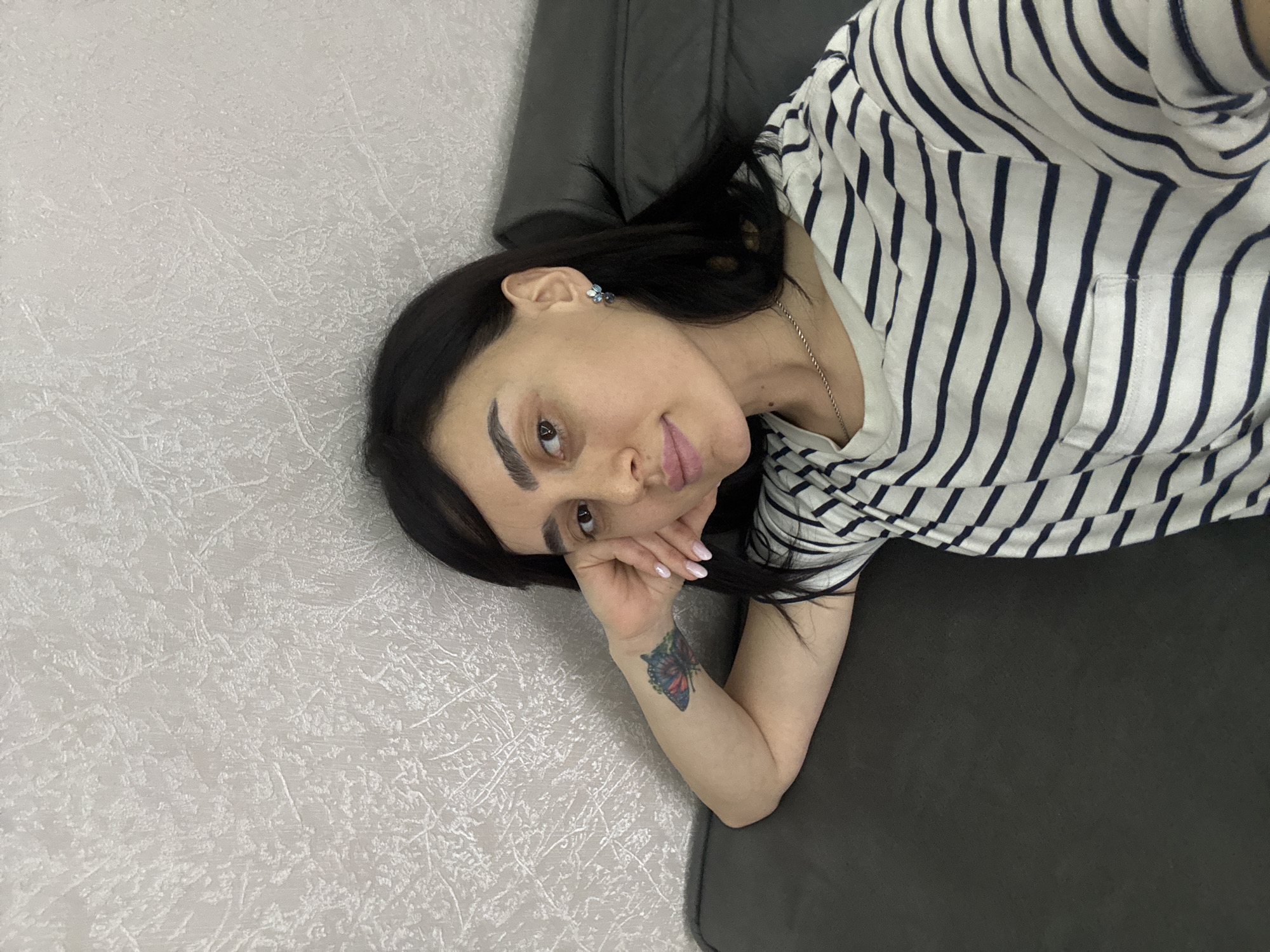 A woman with dark hair is lying on a dark gray sofa in a room with off-white textured walls. She wears a white and dark blue striped T-shirt and an earring. A butterfly tattoo is visible on her left forearm. She rests her head on her left hand and looks at the camera. The location is unknown and cannot be determined from the image.