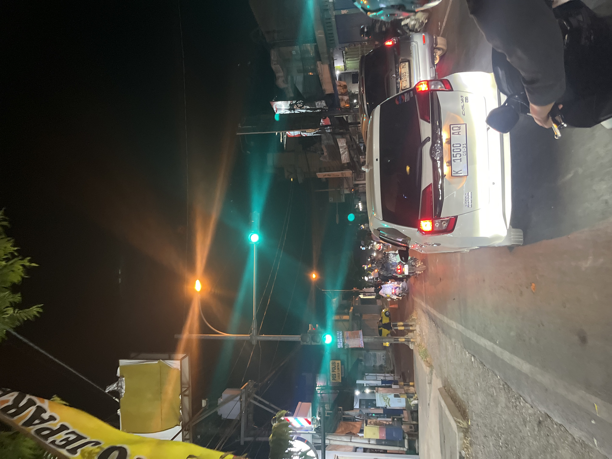 The image depicts a traffic scene at night in the city of Kalinyamatan, Indonesia. A white car with license plate "K 1500 AQ" is in the foreground, its brake lights illuminated. To the right of the white car, a silver car with license plate "K 1985 MQ" is visible. A person is riding a motorcycle behind the white car, with their hands on the handlebars. Several other motorcycles with riders are also present in the traffic. The road is lined with buildings and streetlights. A traffic light is visible, showing a green signal. A sign for "POS POLISI LALU LINTAS" (Traffic Police Post) is mounted on a pole. The overall atmosphere is that of a busy urban street at night.