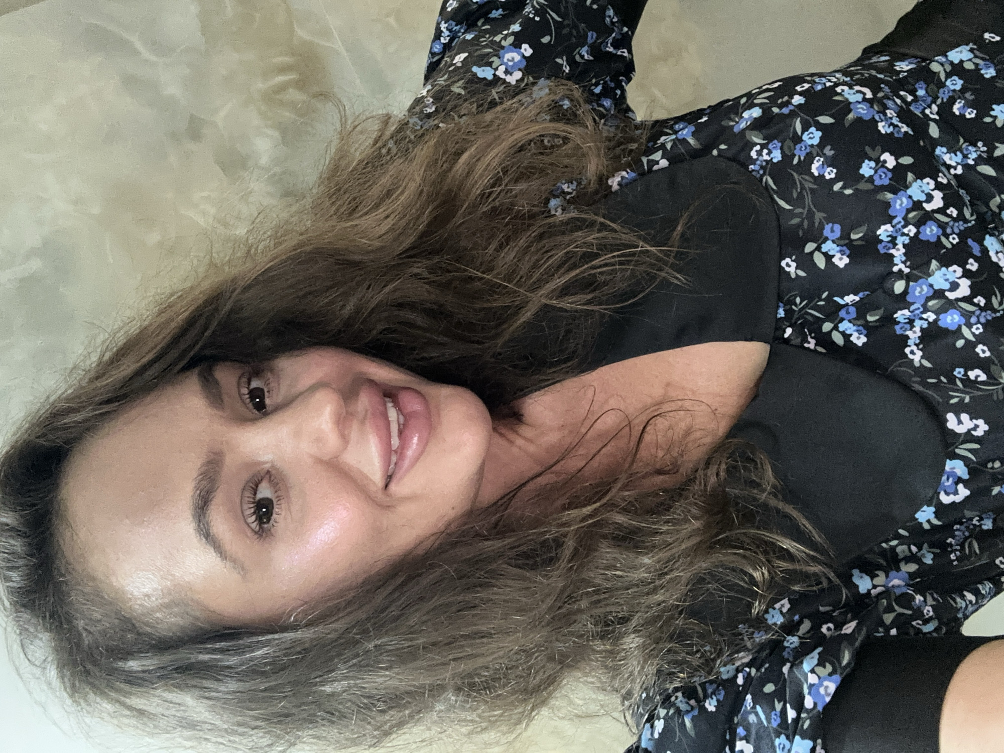 A woman with long, wavy brown hair smiles at the camera. She is wearing a black dress with a blue floral pattern and a black collar. The background is a textured, beige wall. The image is a close-up of her face and upper body.