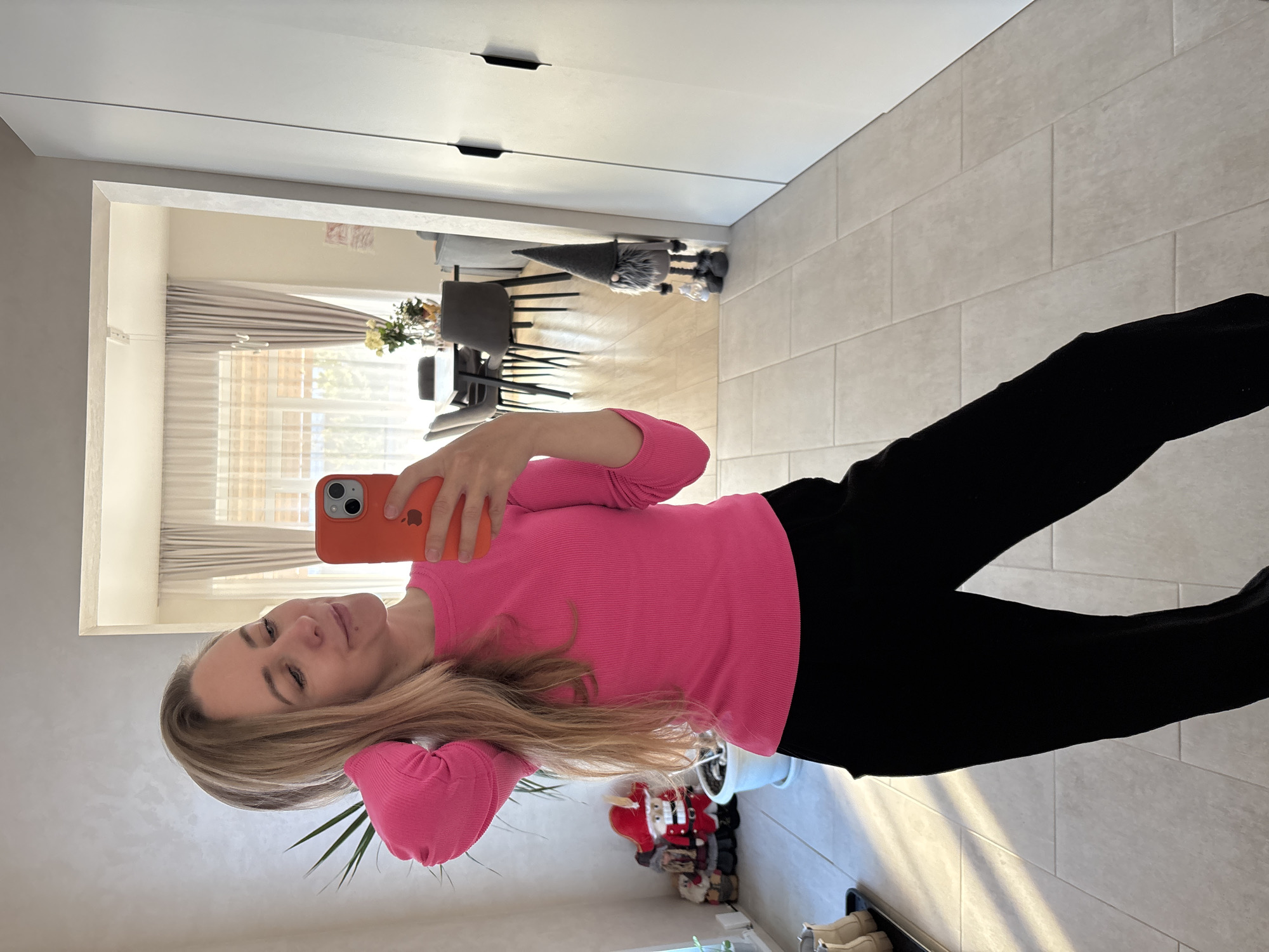 A woman with long blonde hair and a pink long-sleeved shirt is taking a selfie with an orange iPhone in what appears to be an interior space with tiled floors. She is wearing black pants. The room includes a view of a dining area with chairs and a table, visible through a doorway. Other objects in the room include a plant, Christmas decorations, and a gnome figurine. The location is likely a home. The photograph does not provide conclusive evidence to confirm its location in Chaikovsky, Russia.