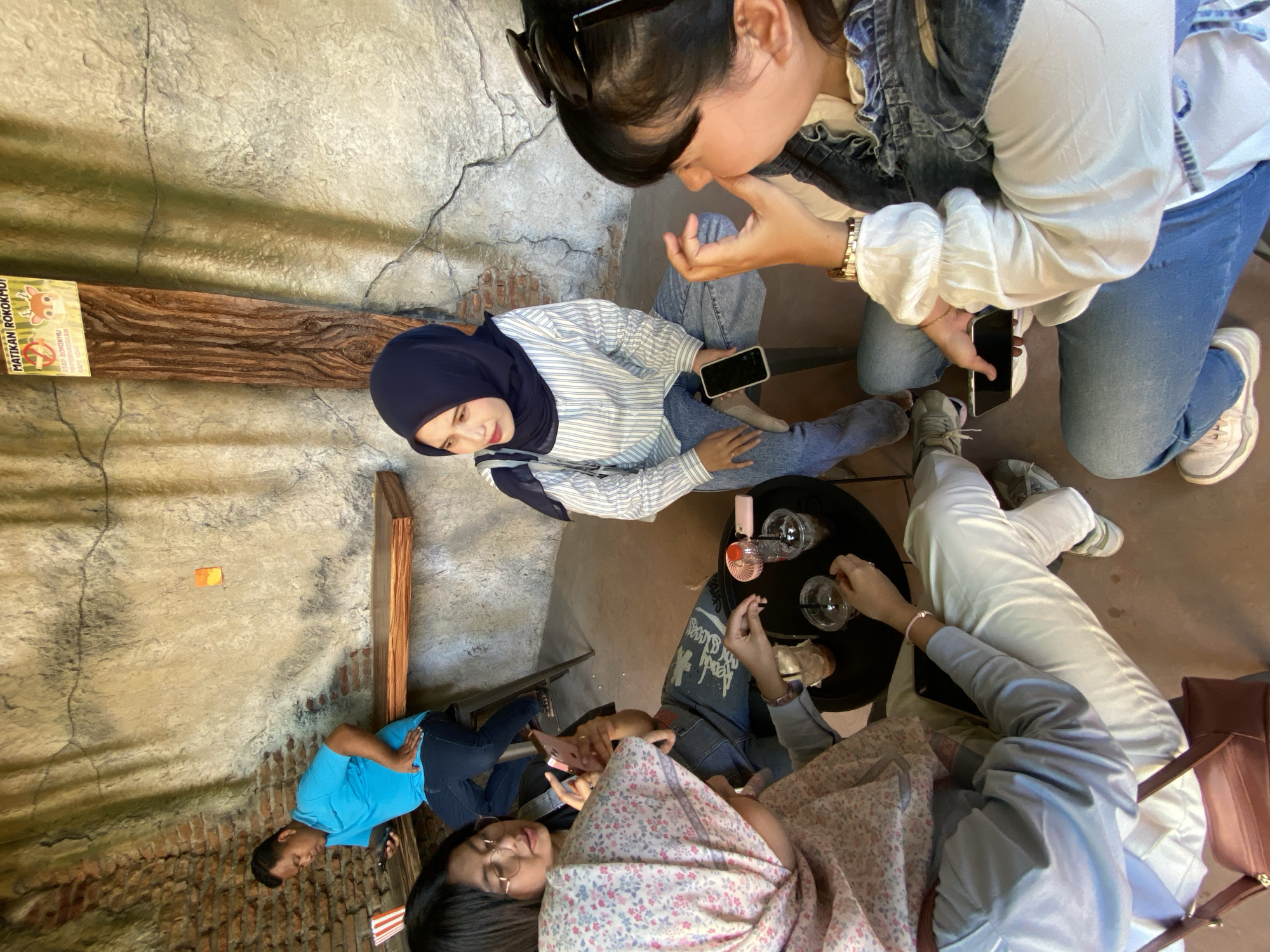 The scene captured appears to be an outdoor seating area in Mangli, Indonesia, characterized by weathered brickwork and faux-naturalistic wall textures. A group of individuals is seated around a low, circular black table holding drinking glasses.
A woman wearing a navy hijab, a horizontally striped shirt, and jeans holds a smartphone and is engaged in conversation with another woman wearing sunglasses on her head, a white blouse, a denim jacket, and jeans. Another individual is wearing a blue shirt and jeans and looking at a phone. At least one more woman is present, wearing a floral-patterned top. Their feet are visible. The group appears to be interacting and socializing.
Attached to a brick wall is a sign in Indonesian, and a small orange rectangle is affixed to the stone wall.
