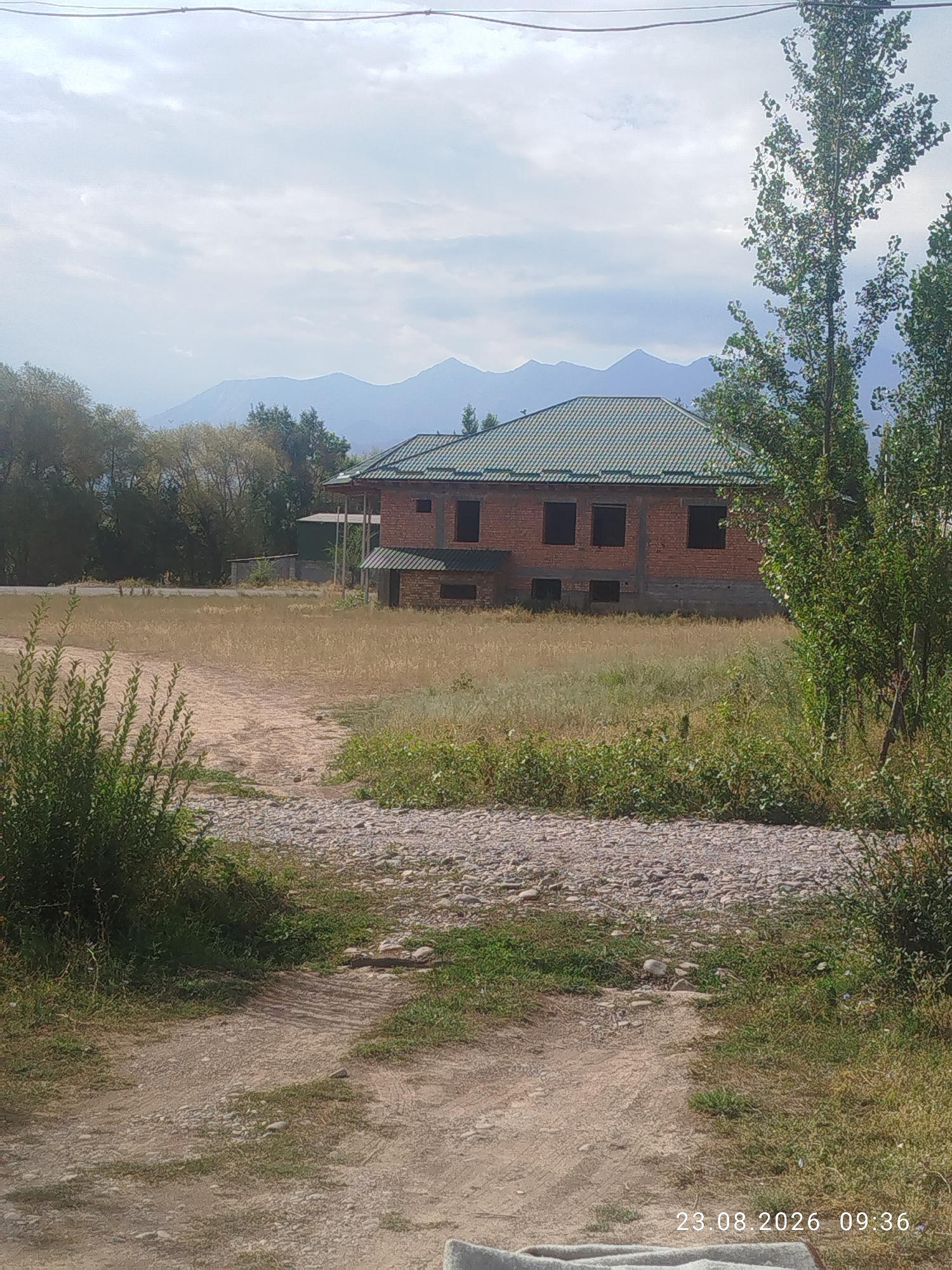 A wide outdoor shot captures a partly constructed brick house with a green tiled roof and dark, empty window frames. The house is situated in a rural area with dry, light-colored grass and brush covering the ground. A dirt path, interspersed with stones, leads towards the viewer from the foreground. In the background, a line of trees and distant mountains are visible under a cloudy sky. The date stamp "23.08.2026 09:36" is visible in the lower right corner, suggesting this image was taken in or around Don-Aryk, Kyrgyzstan.