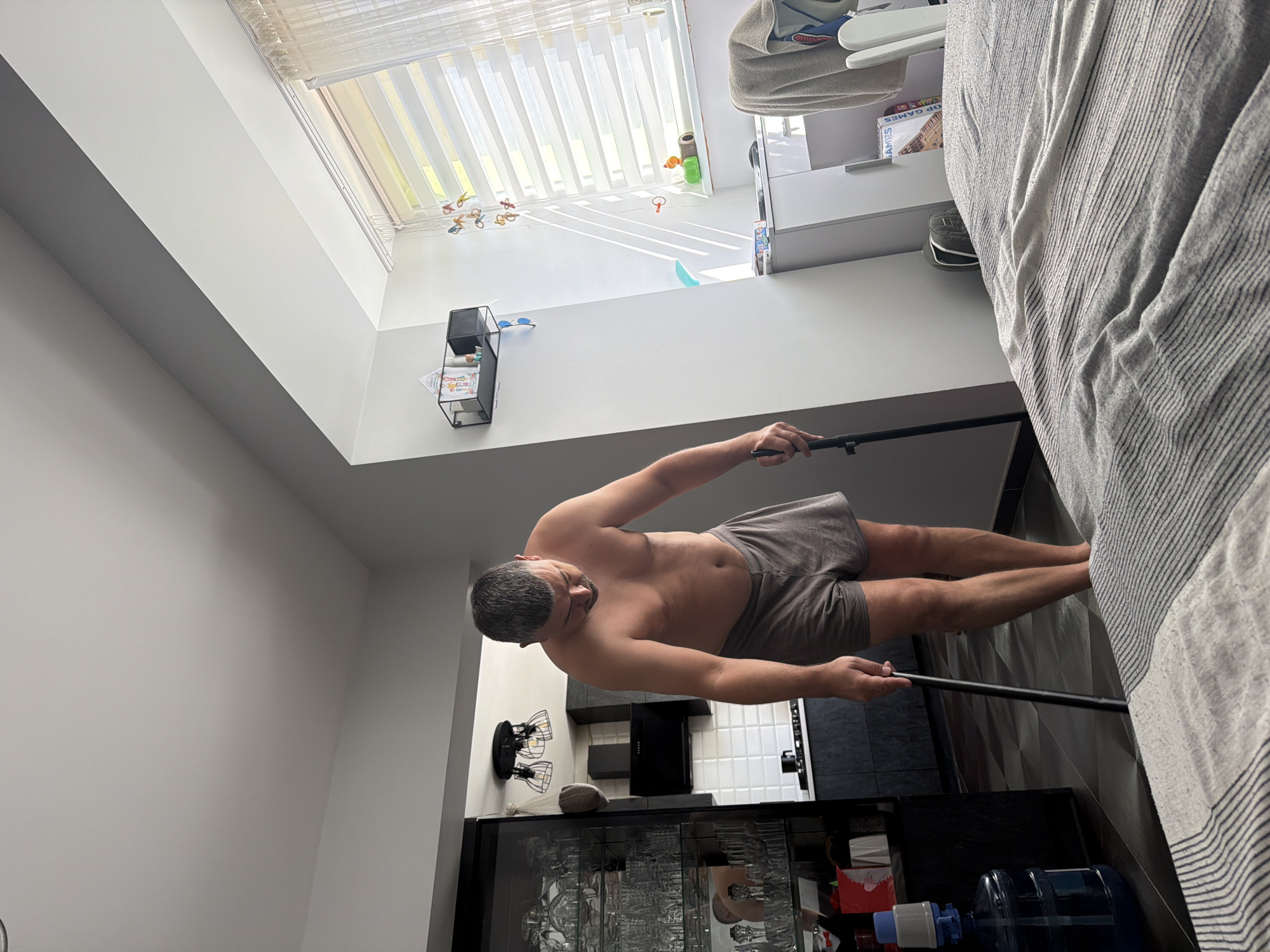 This media file shows a shirtless man in gray boxer briefs standing in what appears to be a kitchen or dining area, holding a long, dark pole. The man is facing away from the camera, looking towards a large window or skylight with vertical blinds. Sunlight streams through the blinds, creating striped shadows on the floor. The room is minimalist, with white walls and dark cabinetry. A large water cooler jug is visible in the lower right corner of the frame, next to a shelf with various items. There is a black cabinet with glass doors that reflect the scene. The image is taken from a low angle, with the perspective rotated so the man appears to be standing on the ceiling.
The man has short, graying hair and a muscular build. His body language suggests he might be engaged in some form of exercise or is holding the pole for support. The overall mood of the image is casual and domestic. The lighting suggests it could be daytime. There is no visible text in the image.