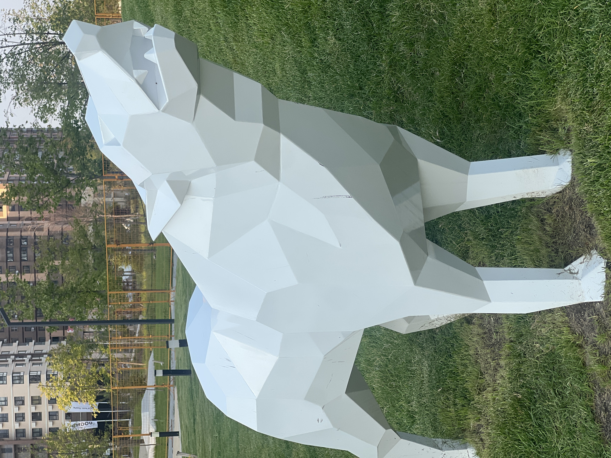 A polygonal white wolf sculpture stands on a grassy incline in a park-like setting. The sculpture is stylized, with sharp geometric shapes forming its body and head. The wolf appears to be howling or snarling, with its mouth open. In the background, there are modern apartment buildings and trees, with a yellow fence visible further back. The ground is covered in green grass. The image was taken in Krasnodar, Russia.