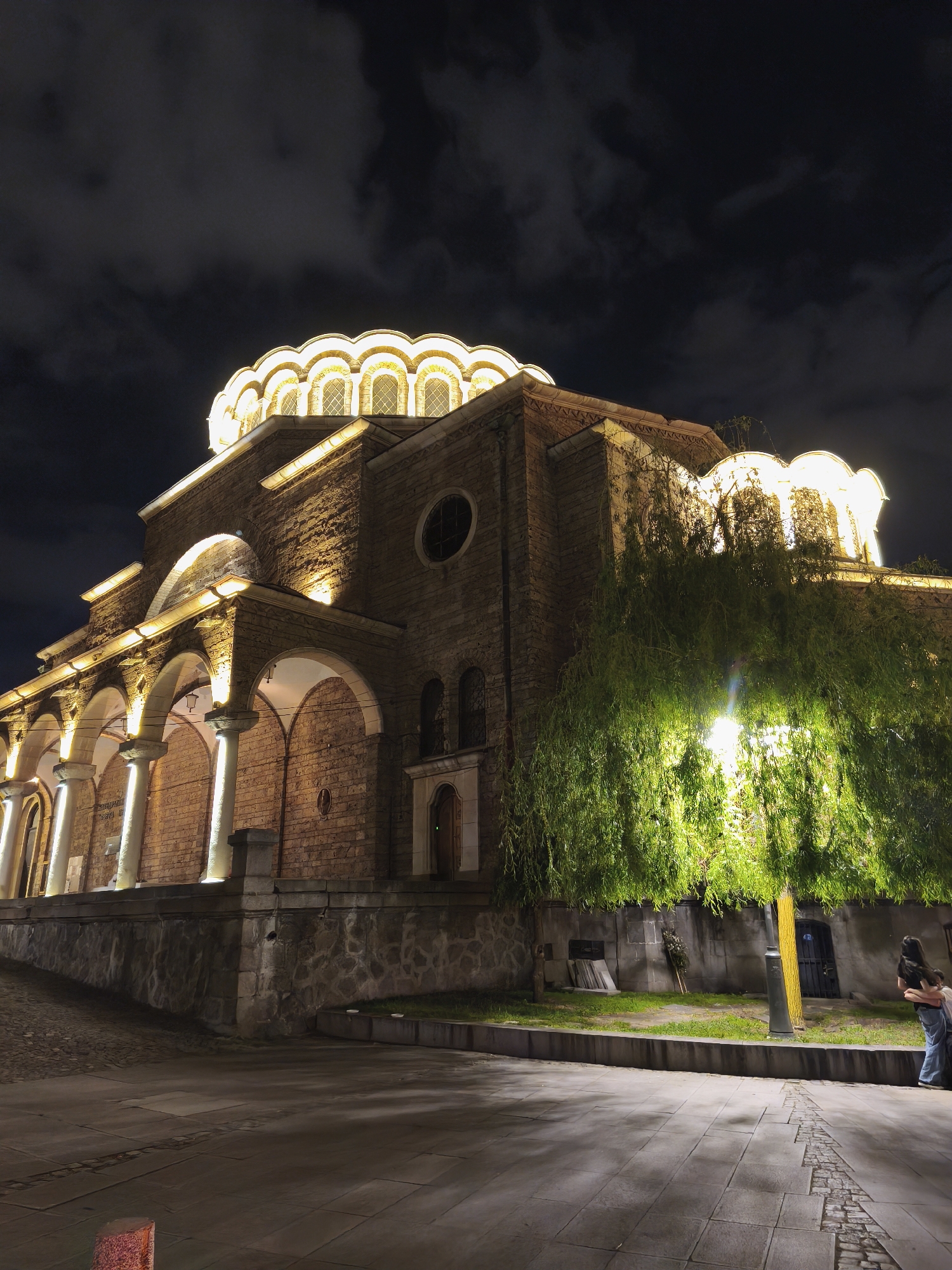 Captured at night, the image features the Church of St. Sophia in Sofia, Bulgaria. The building is made of stone, illuminated with artificial lights, and topped with a dome. To the right of the church, a tree obscures the view with its dense foliage. A street lamp is positioned behind the tree. A person is on the sidewalk by the tree. The ground is paved, and the sky is dark with clouds.