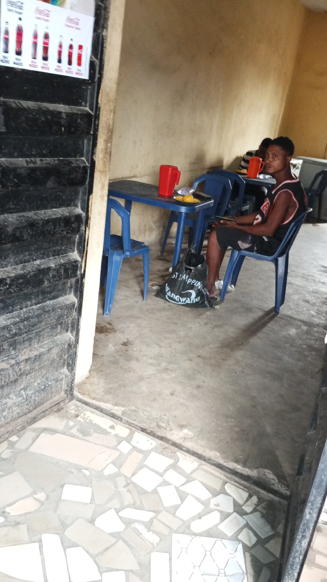 The image depicts the interior of a modest restaurant, possibly in Lokoja, Nigeria. A young man wearing a basketball jersey sits at a blue plastic table with a plate of food and a red pitcher. Another person sits behind him at another table, partially visible. A black shopping bag is on the floor near the table. The floor is tiled in a mosaic pattern, and a Coca-Cola advertisement is attached to the dark stairs on the left. The walls are painted in a faded yellow tone.