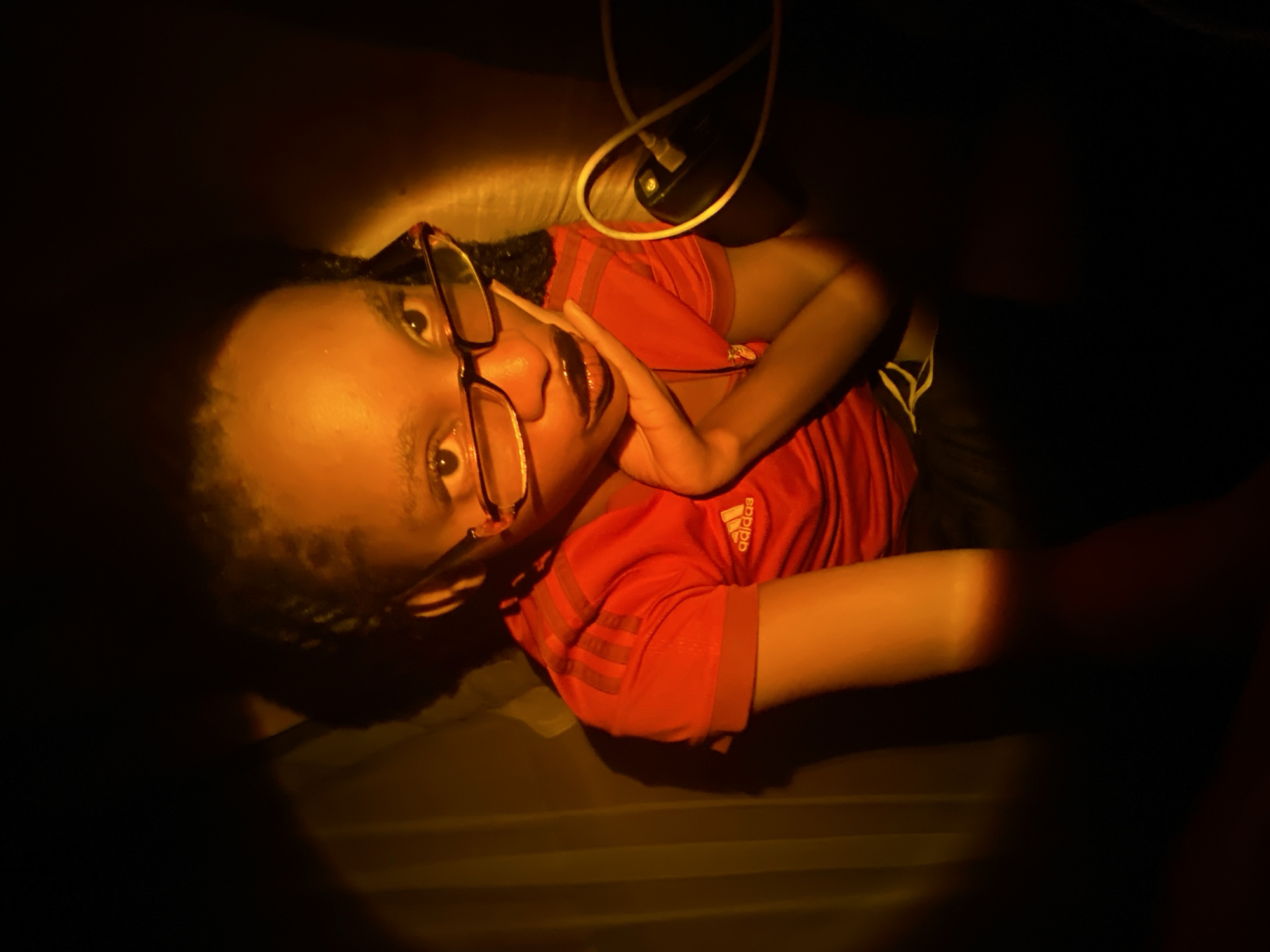 A person with dark skin wearing glasses and an orange t-shirt is lying down. Their right hand is raised near their face. There is a white wire and a black object above them. The scene is dimly lit. The location is Unknown, Unknown.